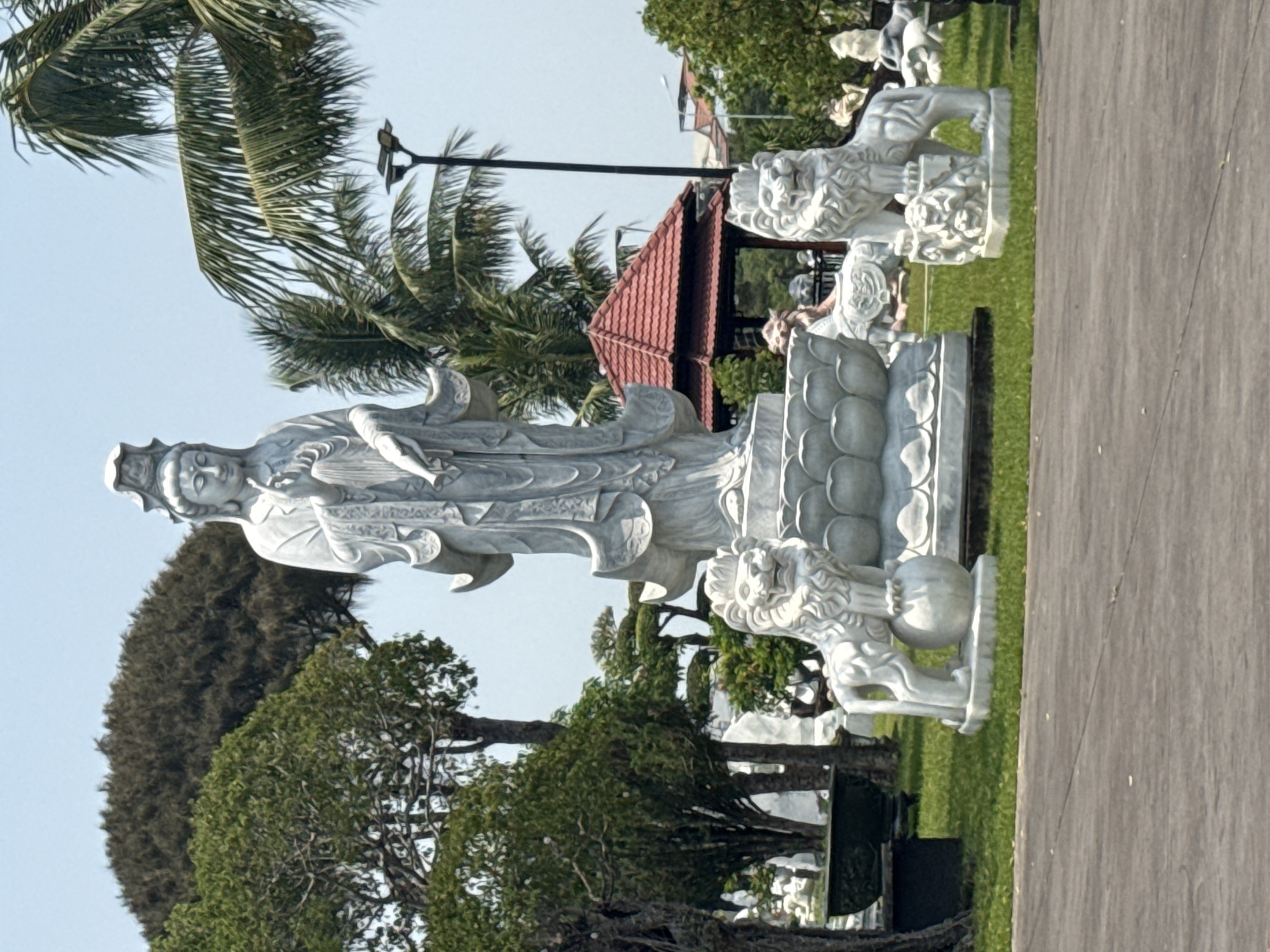 A serene, vertical-oriented photograph presents a marble sculpture of a deity, possibly Quan Yin, a bodhisattva associated with compassion, located in what appears to be a park setting, likely in Đà Nẵng, Vietnam. The sculpture, exhibiting intricate details, is positioned on a pedestal flanked by two marble lion statues, traditional guardians in East Asian cultures. The background features lush greenery, including various trees and palms, against a clear sky. A structure with a red, tiled roof can be observed behind the sculpture. A pathway stretches along the right side of the frame.