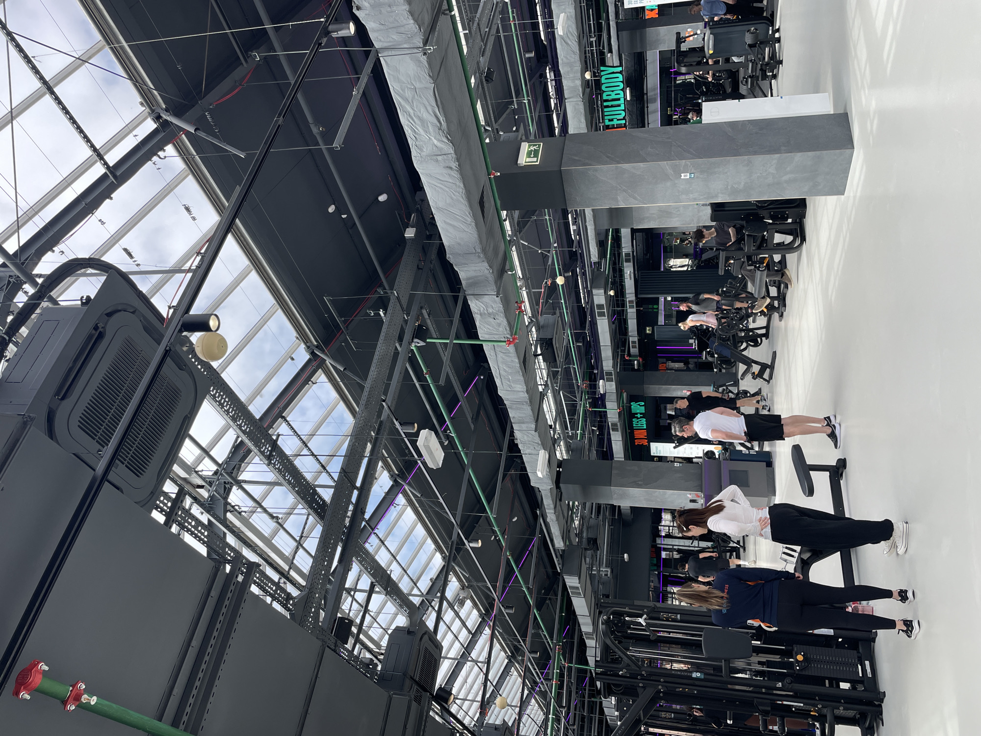 The image depicts the interior of a modern fitness center in Kazan, Russia. The location is identifiable by the "Fullbody" and "30 Minutes Legs and Hips" signage visible in the background. The facility features a high, exposed ceiling with a network of structural steel beams and transparent panels revealing a view of the sky. Multiple people are engaged in various exercises on different fitness machines. Some are using weight machines, while others are positioned for what appears to be stretching or floor exercises. The color palette is predominantly dark, with gray and black equipment contrasting with a white floor. Overhead lighting and ventilation systems are visible.