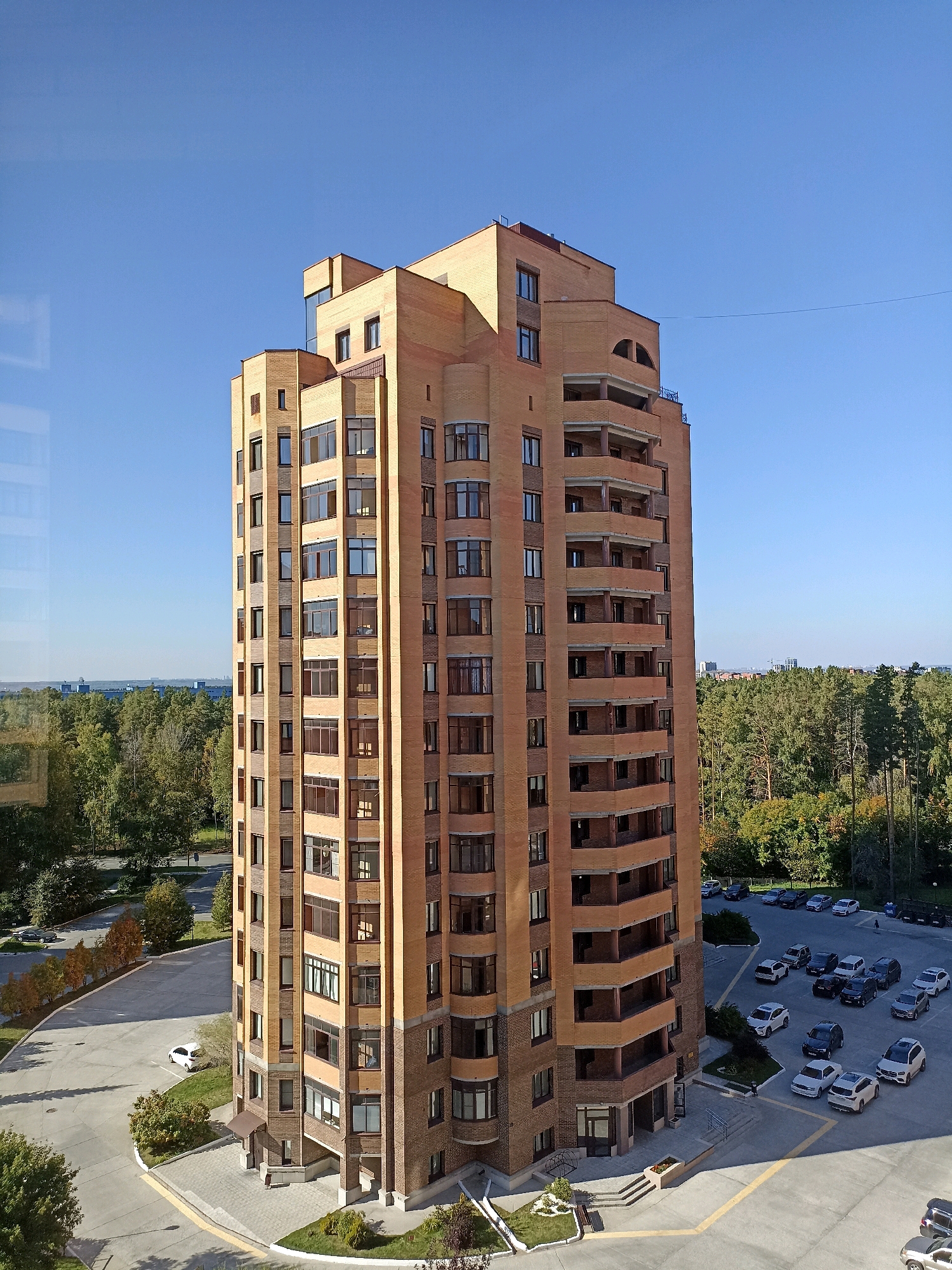 A tall, modern, brick apartment building with multiple balconies and many windows stands in the center of the frame against a clear blue sky. The building is predominantly orange-brown brick with dark-framed windows and beige balconies. To the left of the building, a parking lot with a few cars is visible, including a white car. Further to the left, beyond the parking lot, is a dense area of green trees and some autumnal foliage. The background shows a line of trees and more distant buildings. To the right of the apartment building, another parking lot is filled with numerous cars of various colors and types. Beyond the parking lot and trees on the right, there are hints of more distant urban structures and vegetation. The lighting suggests it is daytime, likely late morning or early afternoon, given the angle of the shadows cast by the building. The scene appears to be a residential area in Novosibirsk, Russia, possibly a modern development on the outskirts of the city given the large expanse of trees.