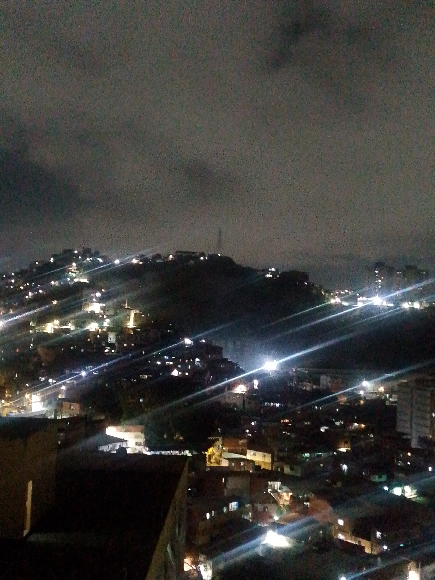The image shows a nighttime view of Caracas, Venezuela. It is captured from an elevated position, likely a high-rise building, overlooking a densely populated urban area. The environment is dominated by a dark and cloudy sky.
The city is characterized by hills covered with buildings, illuminated by scattered lights, creating a constellation-like pattern. A cell phone tower is visible at the top of the tallest hill. The lower parts of the city feature a mix of residential and commercial structures, also illuminated by lights. Streaks of light from vehicle headlights create bright lines across the scene. The scene conveys an impression of urban density and nighttime activity in the city. No people are visible in the image.