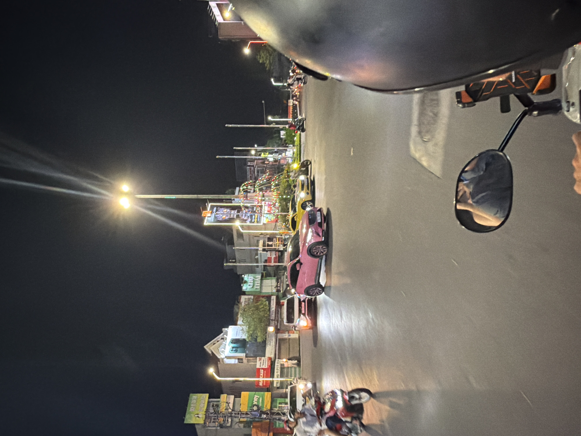 The image captures a detailed night street scene in Tay Nha Trang Ward, Vietnam, from the perspective of someone on a motorbike. In the immediate foreground on the right, a portion of a dark grey motorbike helmet is visible. A rearview mirror attached to the motorbike reflects the upper torso and arm of an individual wearing a blue short-sleeved shirt. Below the mirror, a hand is resting, and a black and orange phone holder is affixed to the motorbike's handlebar area.
The road surface, smooth and grey, occupies the lower and central portions of the image, reflecting ambient light from streetlights and vehicle headlights.
The midground features active urban traffic. On the left, a person in a light-colored shirt and helmet is riding a red motorbike away from the camera, with a passenger partially visible behind them. To their right, a white car with illuminated headlights is visible, facing the camera, possibly making a turn or stopped. In the center, a prominent bright pink Mazda crossover SUV (likely a CX-30) is angled towards the right, also with headlights on. Directly behind the pink SUV, a white Mitsubishi Xpander or similar SUV is visible, also facing the camera with illuminated headlights. Further to the right and slightly behind these vehicles, a yellow Chevrolet Camaro sports car is parked or moving slowly to the left, with a person on a yellow motorbike in front of it. Several other motorbikes and cars are discernible further down the road, creating a sense of a busy thoroughfare.
The background is dominated by multi-story commercial buildings lining the street. Numerous illuminated signs, predominantly displaying Vietnamese text, are affixed to these structures. Notable signs include a large green billboard on the far left depicting a woman and text like "TIẾN VŨ," and other signs with words such as "money," "AL CÁC," "THÁI," "CÁCH," and "CHỢ THÁI." Bright streetlights with multiple lamps punctuate the scene, casting strong light rays into the dark night sky. In the central background, decorative string lights, appearing as colorful dots, are draped between poles, contributing to the urban nighttime ambiance.