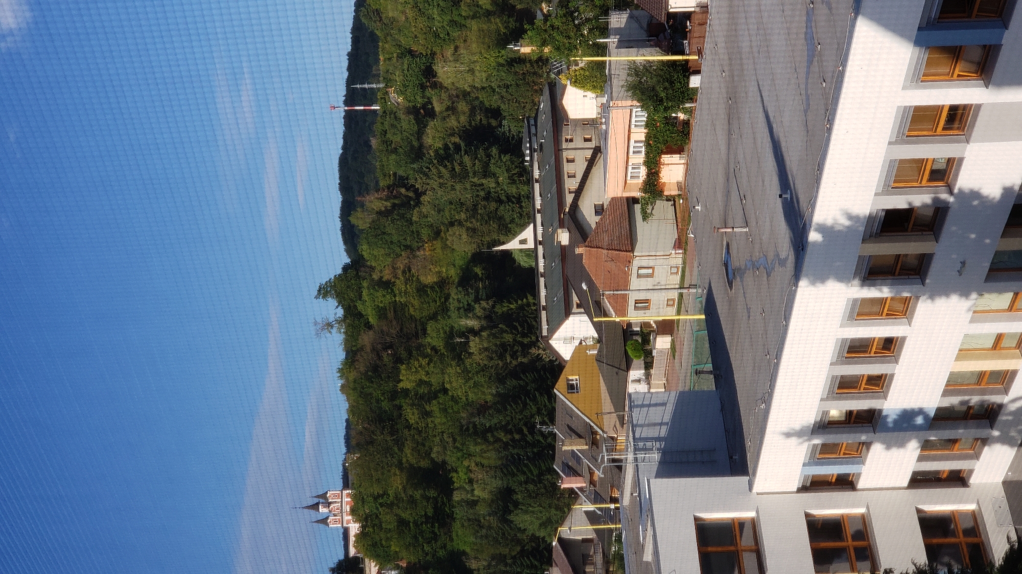 The image shows a view of a residential area with several buildings and a wooded hill in the background, under a clear blue sky. In the foreground, a white building with multiple windows is visible. Behind it, there are several houses with tiled roofs, some of which are yellow or orange. A prominent building with a white facade and dark roof, possibly a school or institutional building, is also present. The hill behind the houses is covered with dense green trees. A church with spires is situated on the left side of the hill. On the right side of the hill, a red and white communication tower is visible. The scene appears to be captured from an elevated perspective, looking down on the neighborhood. The presence of the church, the communication tower, and the general architectural style suggest a location in Eastern Slovakia, potentially within or near the city of Prešov. The overall ambiance is peaceful and serene, with the sunlight casting shadows on the buildings.