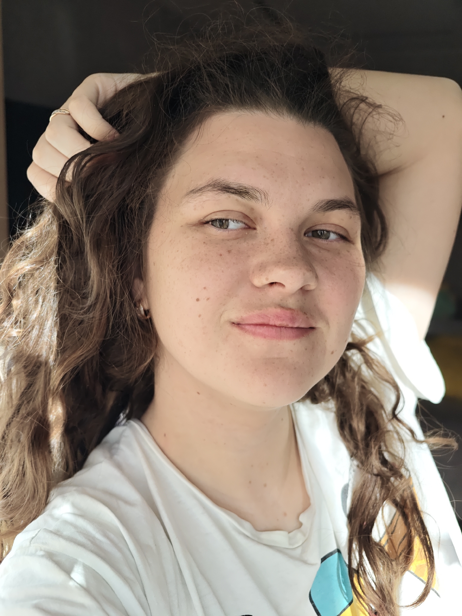 A young woman with long, curly dark brown hair is depicted in a close-up, chest-up portrait taken in Chaikovsky, Russia. Her fair skin is adorned with numerous freckles across her nose and cheeks. She looks directly at the viewer with light green or hazel eyes and a gentle, subtle smile. Her lips are naturally pink and full.
Her left hand is raised, with her arm bent and reaching over her head, as she casually runs her fingers through her hair on the top left side of her head. A simple ring is visible on her left ring finger. A small, round, gold-colored hoop earring can be seen in her right ear. She is wearing a white short-sleeved t-shirt, with a partial design featuring light blue and yellow geometric shapes visible on the lower right portion of the garment.
Strong natural light illuminates her from the left, highlighting her features and creating a warm glow in her hair, suggesting the photograph was taken during the daytime. The background is dark and out of focus, implying an indoor setting. Her relaxed pose and pleasant facial expression convey a sense of calm. There is no visible text in the image.