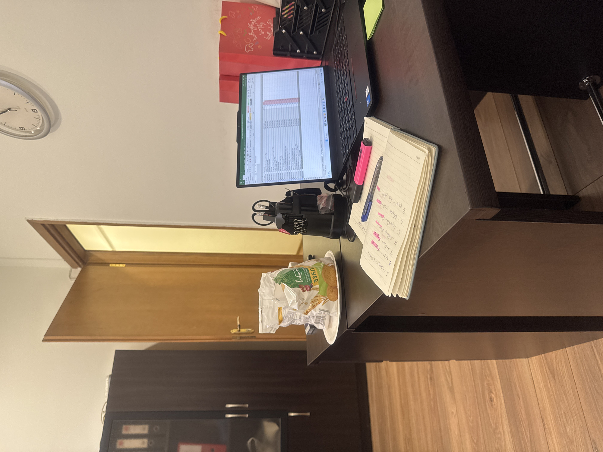 The image captures a desk scene, featuring a laptop displaying a spreadsheet, a notebook with handwritten notes, writing utensils, and a partially consumed bag of "Juice" crackers resting on a plate. The desk is a dark wood color. In the background, a closed wooden door is visible along with a dark cabinet with silver handles. The scene is likely indoors. While the image provides no explicit information confirming its location in Tbilisi, Georgia, it offers no reason to disbelieve that claim. There are no people visible in the image.