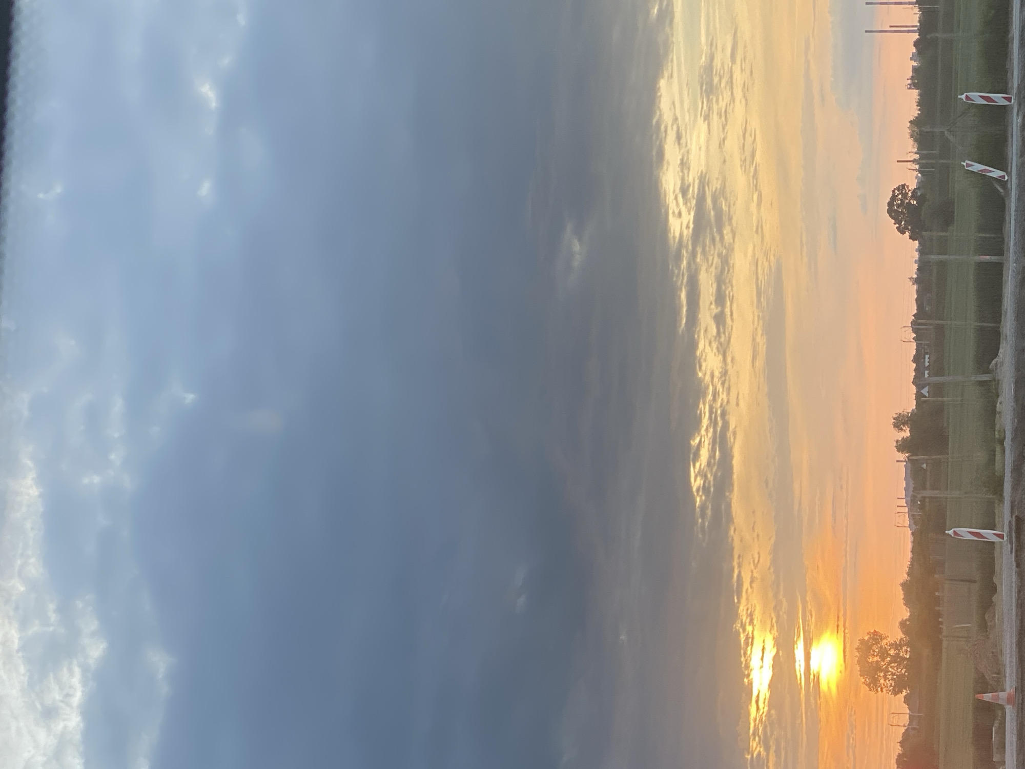 The image depicts a landscape at dusk, likely in Podłęże, Poland. A vibrant sunset casts an orange and yellow glow across the sky, illuminating scattered clouds. In the foreground, a construction zone is visible, with a safety cone and striped bollards. Beyond the construction site, a field stretches towards a treeline, with several industrial smokestacks in the distance. The scene is devoid of people.