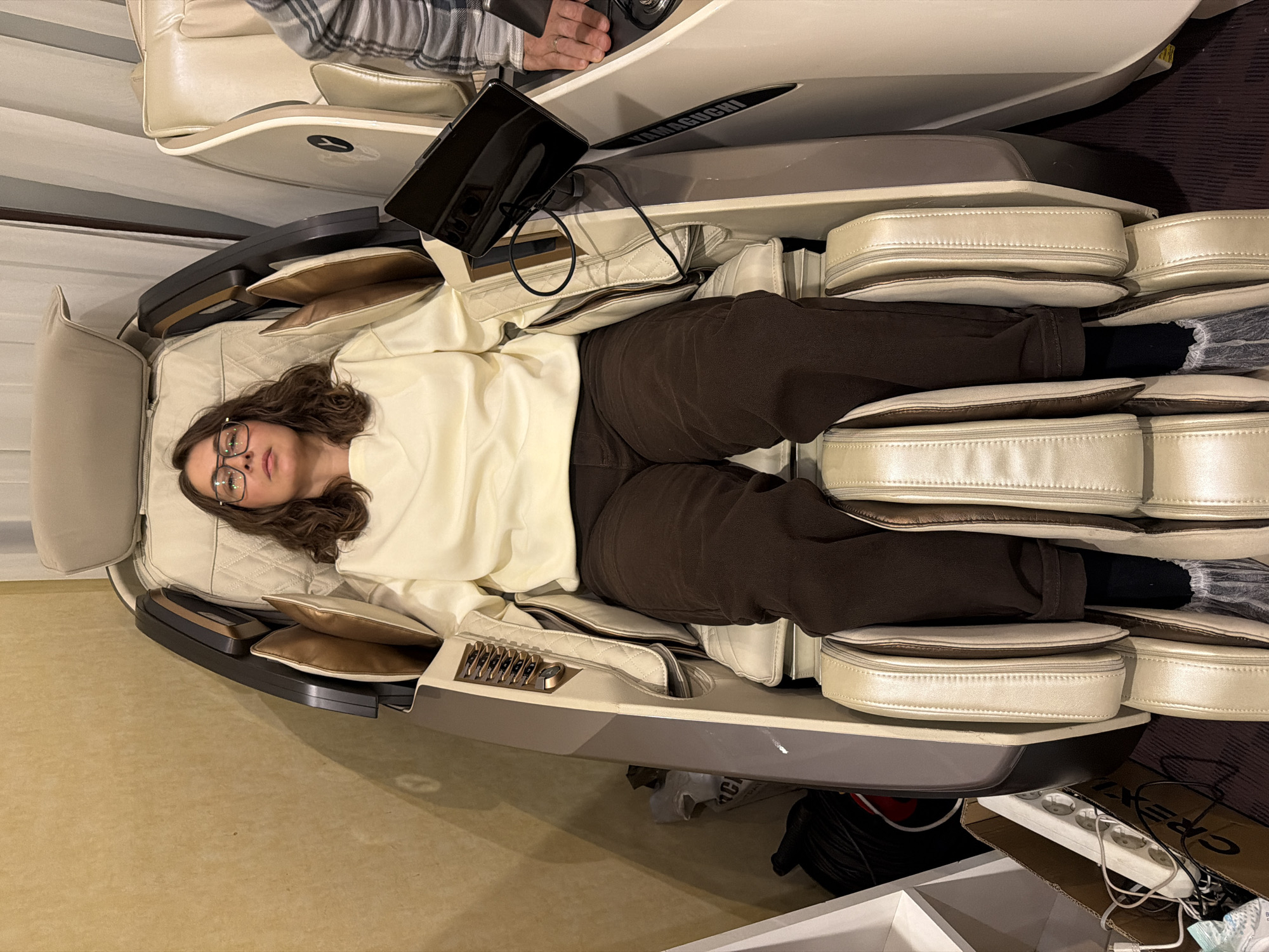 A woman wearing glasses, a cream-colored sweater, and brown pants is lying in a Yamaguchi massage chair. The massage chair is located in what appears to be a room, possibly in a building in Izhevsk, Russia. A tablet device rests on the chair near her head. The chair has a cream and brown color scheme, with multiple layers of padding. The area around the chair contains various items, including a box, cords, and what looks like an electrical power strip. Another person is partially visible near the top-left of the frame. The image is taken from an elevated perspective.
