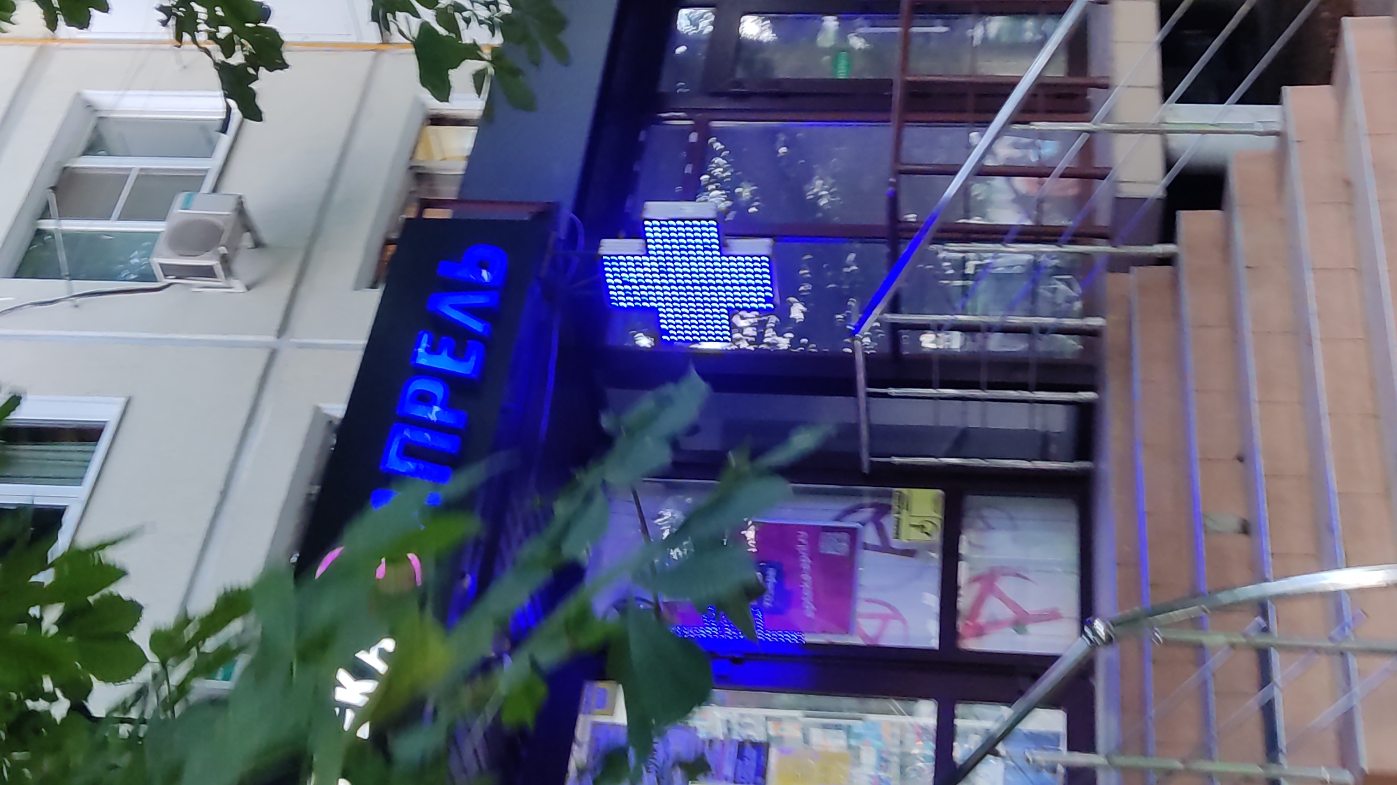 The image shows the facade of a pharmacy with a blue illuminated cross sign and blue neon lettering on a black background. The lettering appears to read "АПТЕКА" (Apteka), meaning pharmacy, followed by another word in blue. In front of the pharmacy, there are stairs with a metal railing leading up to the entrance. Trees with green leaves partially obscure the view of the building. The pharmacy is likely located in the city of Krasnodar, Russia, as indicated by the context provided.