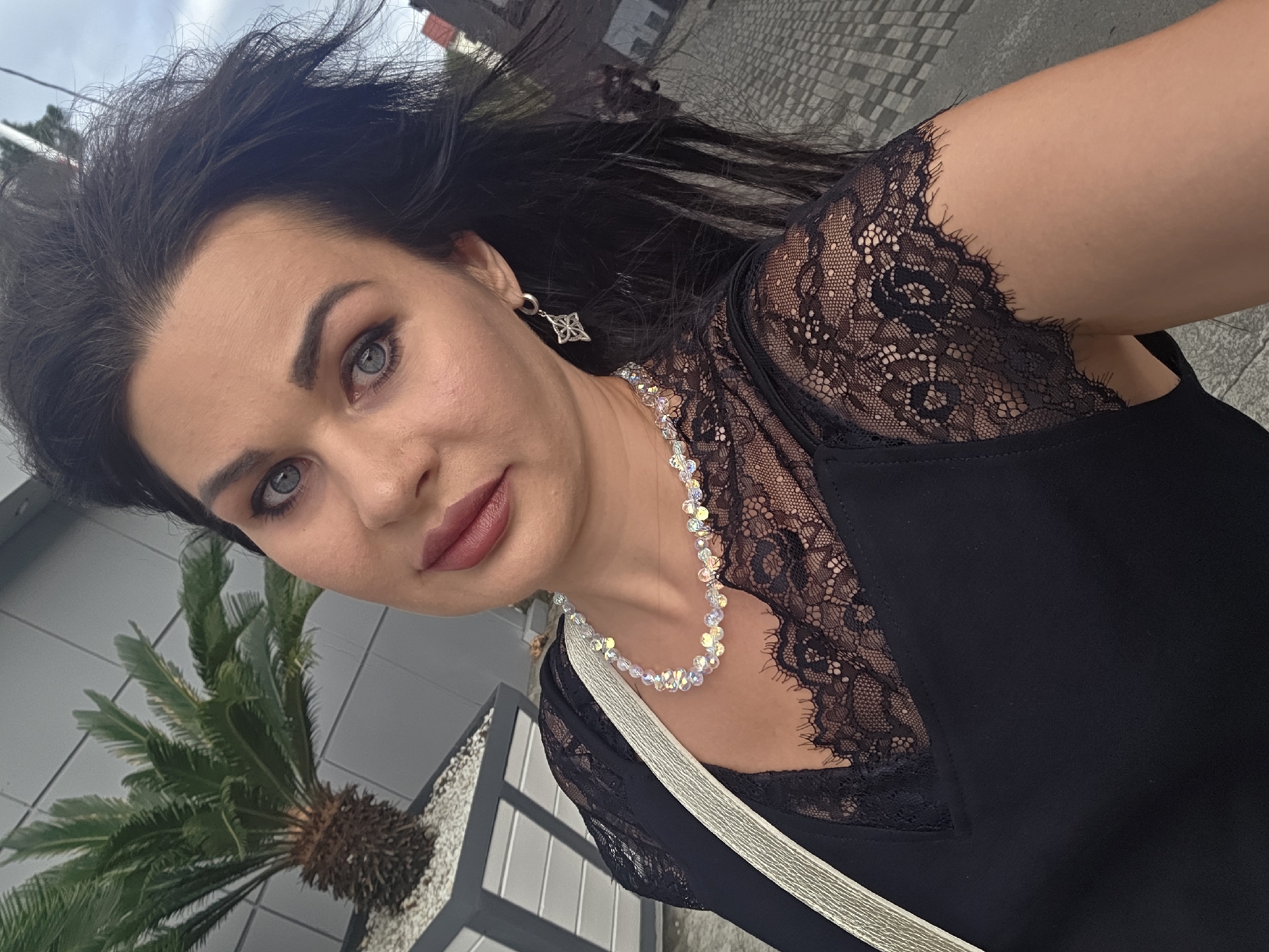 This is a selfie of a woman taken outdoors. She has long, dark hair that is windswept, and she is looking directly at the camera with a neutral expression. She is wearing a black top with lace detailing on the shoulders and neckline, a crystal necklace, and a diamond-shaped earring. A pale gold strap is visible across her chest.
In the background, there is a potted palm plant with green fronds, and a grey, tiled surface. Behind that, there appears to be a building with windows and a gray, textured exterior wall. The sky is overcast. The overall impression is of a casual daytime photograph, likely taken in a city or urban environment.