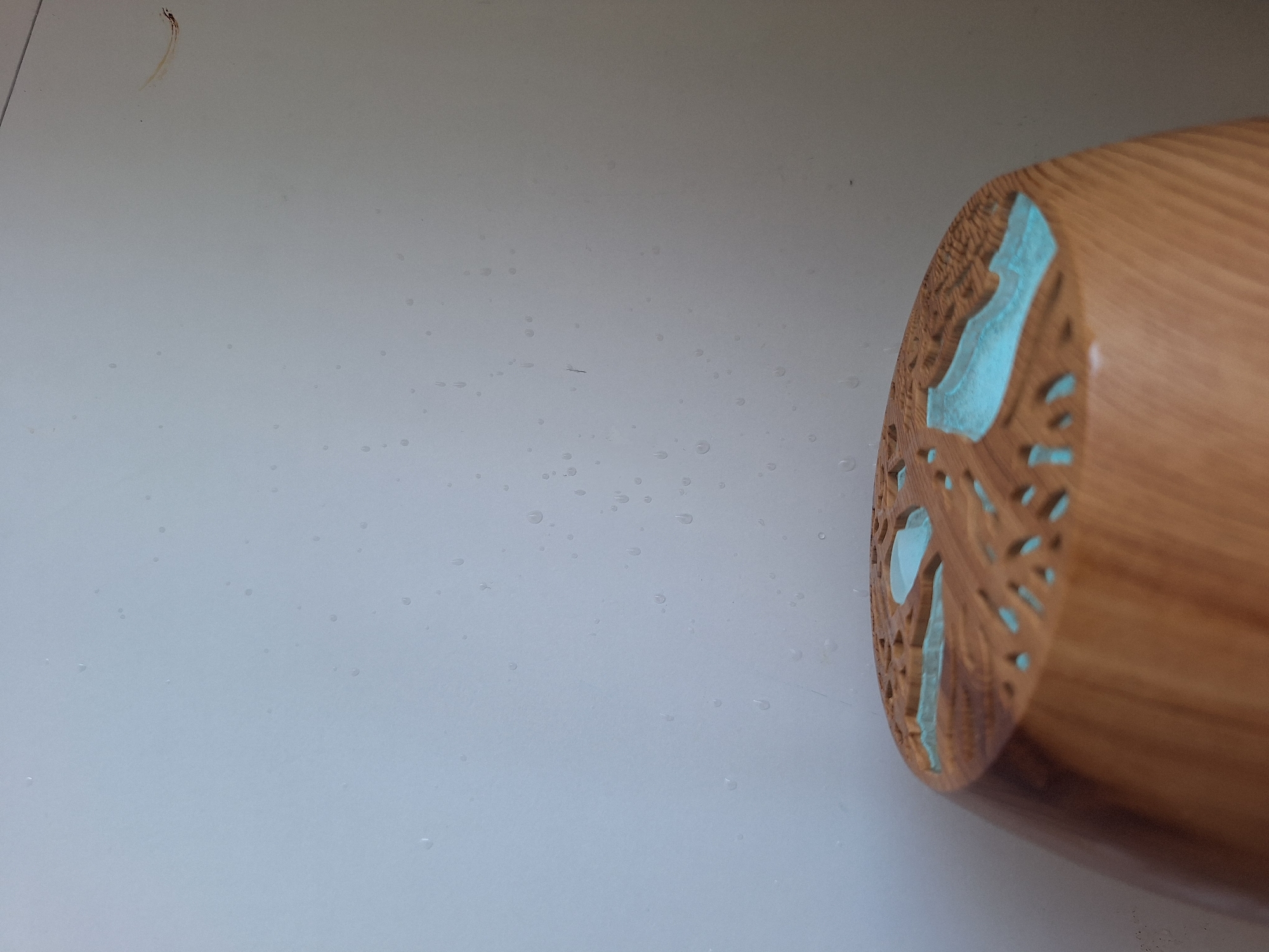 The image shows the top of a wooden diffuser with intricate cutouts filled with a turquoise resin. The diffuser is made of light brown wood with a visible grain. The background is a plain, light-colored wall with several small water droplets or dust specks. There are no people or animals visible in the image. The lighting suggests an indoor setting, possibly near a window, as evidenced by the reflection on the wall. There is no information in the image that allows for the determination of its specific location within Varna, Bulgaria.