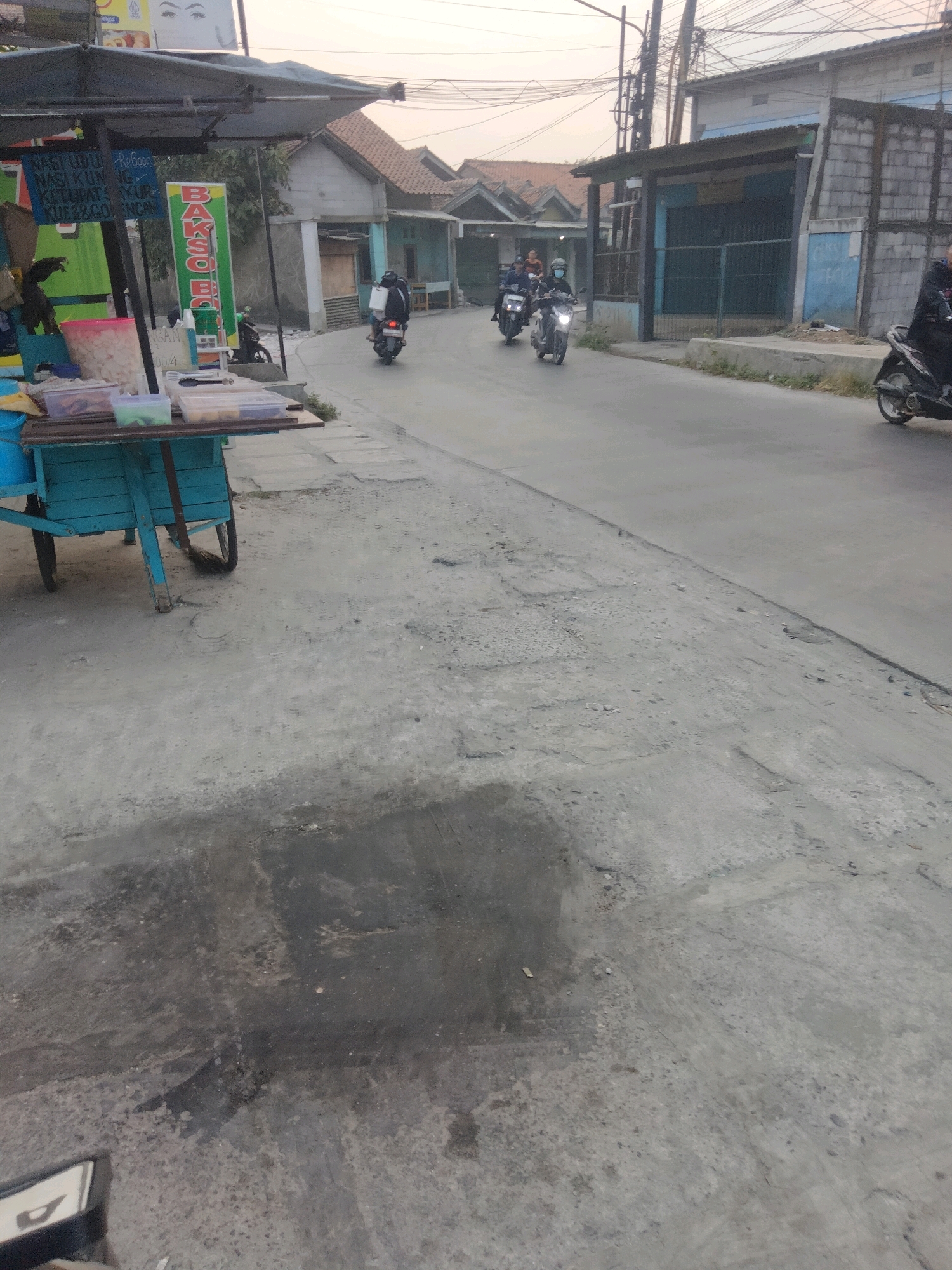 In Pisangan Jaya, Indonesia, an overcast sky looms over a street scene with a food cart and passing motorcycles. A blue and teal food cart, laden with containers of food and snacks, sits on the left side of the image. A vertical sign with green and red text is attached to the cart. Several motorcycles are visible on the asphalt road. One motorcycle, ridden by a person wearing a dark jacket and helmet, is in the foreground on the right. Further down the road, three other motorcycles, with riders also in protective gear, are moving away from the camera. The surrounding environment features buildings with pitched roofs and some with exposed concrete block walls, as well as utility poles and overhead wires. The road surface shows signs of wear and tear, with patches of darker, wet-looking areas.