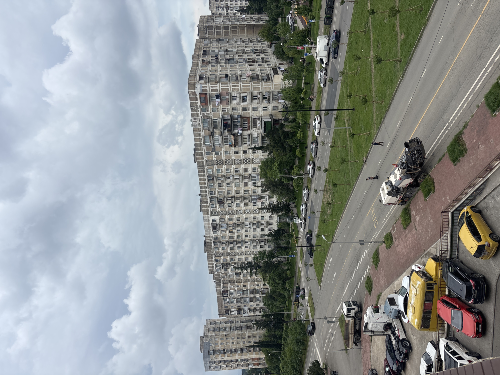 The image shows a view of a street in Tbilisi, Georgia with several tall apartment buildings lining one side. The buildings are concrete and appear to be from the Soviet era. A multi-lane road runs alongside the buildings with cars parked on the side. There are trees and green spaces separating the road from the buildings. Several people are walking on the road. A large piece of construction equipment is visible on the road. The sky is overcast.