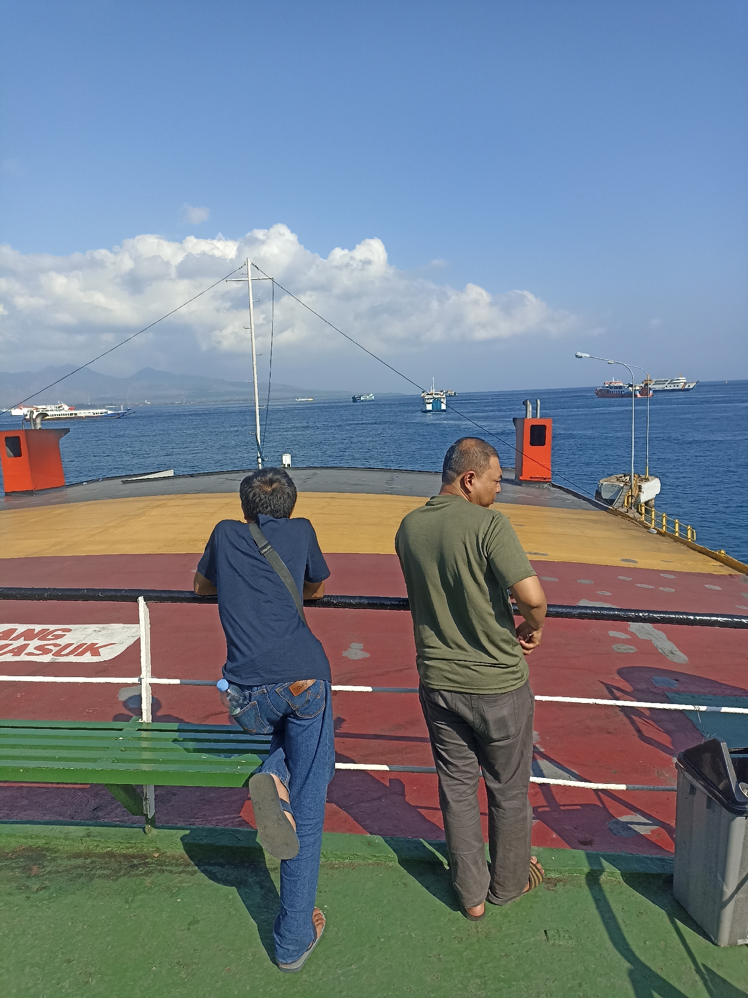 Two men stand on the deck of a ferry, looking out at the ocean. The man on the left wears a dark blue t-shirt and jeans, with a grey strap across his chest. He leans on a railing, his right leg extended. The man on the right wears a green t-shirt and dark pants, with his hands clasped behind his back. The ferry deck is painted red and yellow. In the background, several ships are visible on the blue water, and a hazy coastline with mountains can be seen under a bright blue sky with scattered clouds. The scene appears to be in the waters near Ketapang, Indonesia, a port city known for its ferry connections.