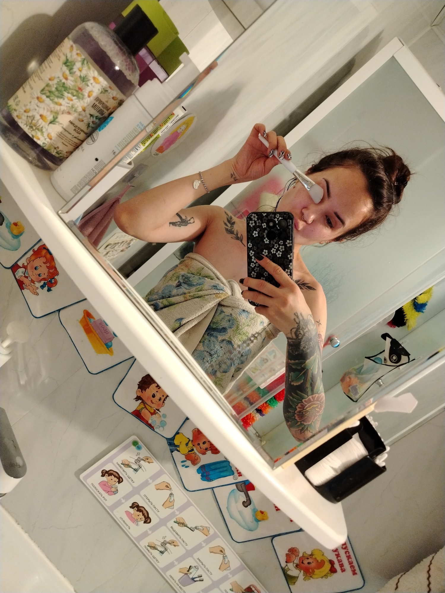 The image shows a woman taking a selfie in a bathroom. She is partially wrapped in a towel and is applying something to her face with a brush. Her phone has a black case with a white floral pattern. Tattoos are visible on her arms and shoulder. The bathroom has various items, including bottles of toiletries and a drawer filled with tissues. Educational mats depicting hand washing and teeth brushing are placed on the floor. One of the bottles has Russian text on it, and a mat on the floor in Russian suggests the scene might be in a Russian-speaking area, potentially the city of Noviy, Russia, as stated in the prompt.