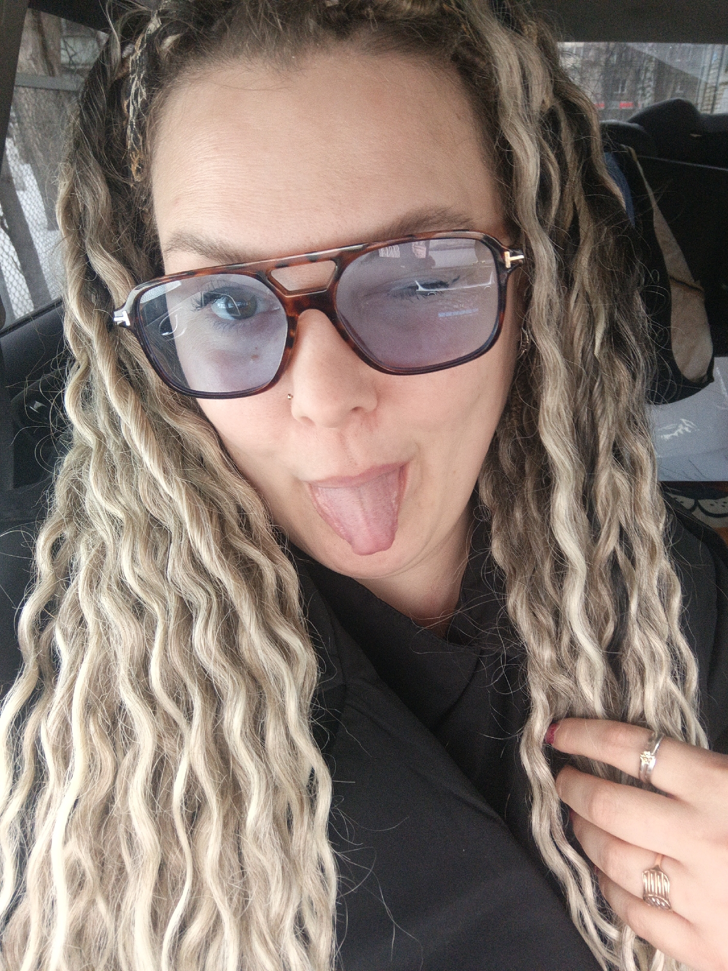 An adult female is depicted in a close-up selfie, positioned inside a vehicle. She has long, wavy hair with blonde highlights and darker roots, partially styled with braids near the scalp. She wears brown tortoise-shell aviator-style sunglasses with purple-tinted lenses, featuring a small silver 'T' emblem on the left temple. Her green eyes are visible through the lenses. A silver stud piercing is present on her left nostril. Her mouth is open, with her tongue extended outward and downward. She is wearing a dark-colored top or jacket. Her left hand is partially visible, resting near her hair, displaying a silver-colored ring with a square clear stone on the index finger and a gold-colored, multi-band ring on the ring finger. Her index fingernail is painted dark red. The blurred background, visible through the vehicle's window and reflected in the sunglasses, indicates an urban environment with indistinct buildings and structures. A precise location within the city of Chaikovsky, Russia, cannot be determined from the image.