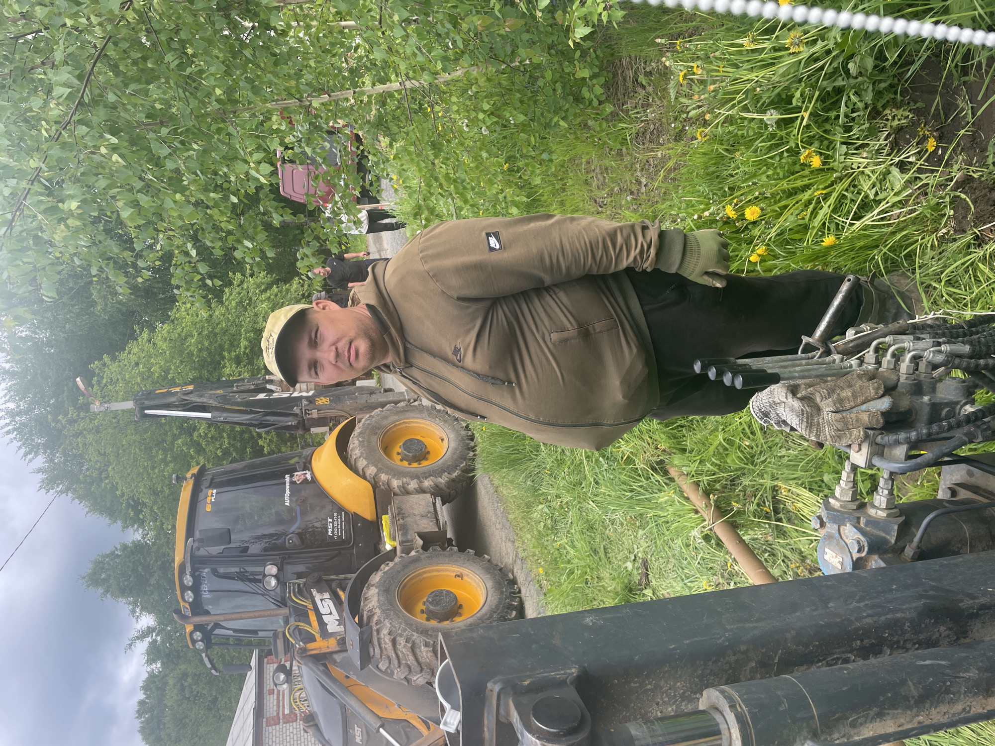 This image depicts a male individual standing in an outdoor setting, likely a work site, in the city of Железнодорожный, Russia. The man, of heavy build and appearing to be in his 30s or 40s, has a neutral expression and looks directly at the camera. He wears a camouflage baseball cap, a dark green Nike hooded sweatshirt with a Nike logo on the sleeve, dark pants, and green work gloves. In the immediate foreground, to the left, are dark-colored hydraulic cylinders and control levers of heavy equipment, upon which a worn work glove rests. A light brown pipe or conduit is visible on the ground amidst green grass and yellow dandelions. Behind the man, a yellow and black MST 655 Plus backhoe loader is parked on a dirt path. The backhoe's cab features "СЕРЕГА" (SEREGA) on its top, "655 PLUS" and "AUTOpowershift" on its side, and "MST" branding with Russian contact details, including a phone number and website (www.mst-ural.ru). Its rear backhoe arm extends upwards. Two other men are visible walking away in the background along the dirt path, past a partially obscured reddish-brown truck. The scene is surrounded by abundant green foliage, including trees and bushes, with a light-colored brick building partially visible on the far left. The sky is overcast.