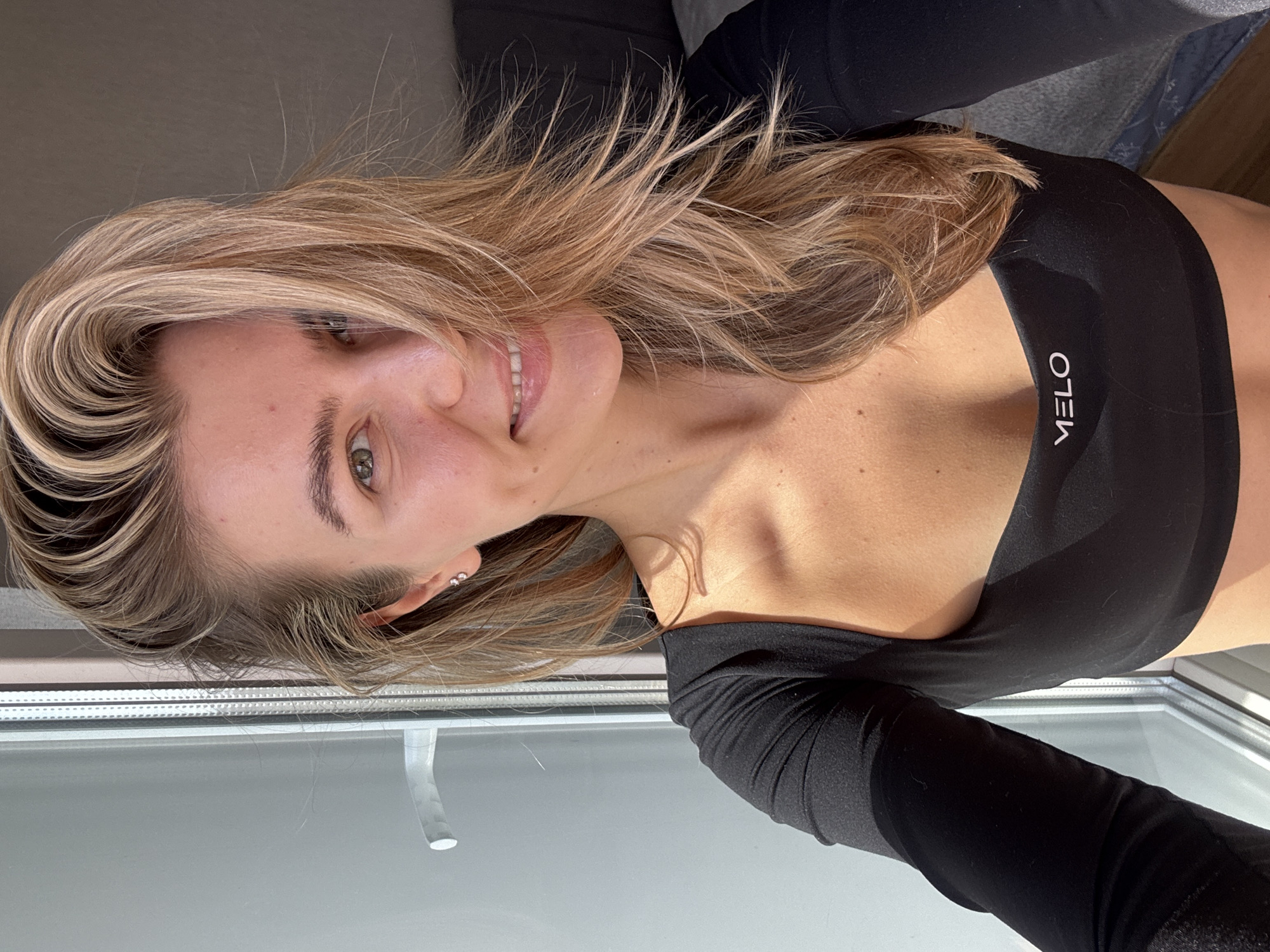 The image displays a head-and-shoulders portrait of a young woman, rotated 90 degrees counter-clockwise. She has long, wavy hair with blonde highlights and light brown undertones, styled to her right side. Her face is turned towards the viewer, and she is smiling gently with her lips slightly parted, revealing white teeth. Her eyes appear green or hazel.
She is wearing a form-fitting, black long-sleeved top or crop top. On the right side of her chest, the brand name "MELO" is visible in white, vertically oriented text. She has small, silver stud earrings in both ears. Her skin appears tanned, with natural light catching highlights on her cheekbones and collarbone.
The lighting is bright and natural, indicating daytime, possibly sunlight streaming from the side, creating distinct highlights and soft shadows across her skin and hair. The background consists of a dark, blurry area behind her head, possibly a piece of furniture, and a lighter, out-of-focus window frame or sill at the bottom, suggesting an indoor setting. The woman's expression conveys happiness and a relaxed demeanor.