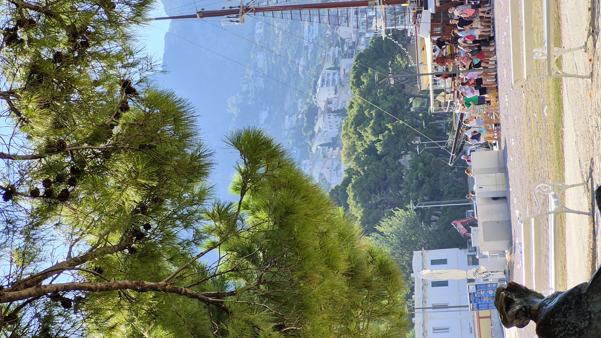 The image captures a bright, sunny day in an outdoor setting with a harbor and hillside town in the background. In the foreground, a statue's head and shoulder are visible on the left, partially obscuring a white park bench. Another white park bench is situated to the right. Pine branches and needles hang from the top of the frame, casting shadows.
In the middle ground, a line of people, appearing to be tourists, stands along a waterfront promenade. They are dressed in casual summer clothing. Behind them, several boats are docked, including a large, traditional sailing vessel with masts and rigging. A white building with green-shuttered windows is also visible on the left. Further in the background, a hillside town with numerous white buildings is spread across a verdant, mountainous landscape. The sky is clear and blue. The ground in the middle ground appears to be a sandy or gravelly area with scattered litter. An excavator is parked near the white building.