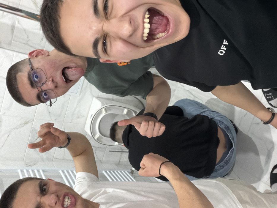 The image depicts four young males in a white-tiled bathroom in Panagyurski kolonii, Bulgaria. Three individuals are prominently positioned in the foreground and mid-ground, making exaggerated facial expressions and gestures towards the camera. The subject in the immediate foreground right wears a black "OFF" t-shirt, with a wide-open mouth, clenched eyes, and a black braided bracelet on the left wrist. To the left, another subject in a white t-shirt shows bared teeth, clenched fists, and a pointed finger, wearing a black wristband and a thin black string bracelet. Between them, slightly behind, a third subject with glasses and a dark green t-shirt sticks his tongue out and gives a "thumbs up" with his right hand, adorned with a black smartwatch. In the background, a fourth male, dressed in a black long-sleeved shirt and light blue jeans, is hunched over an open white toilet bowl, his back to the camera, suggesting illness or distress. The bathroom features white marble-effect wall tiles, a white floor, and a white heated towel rail on the left.