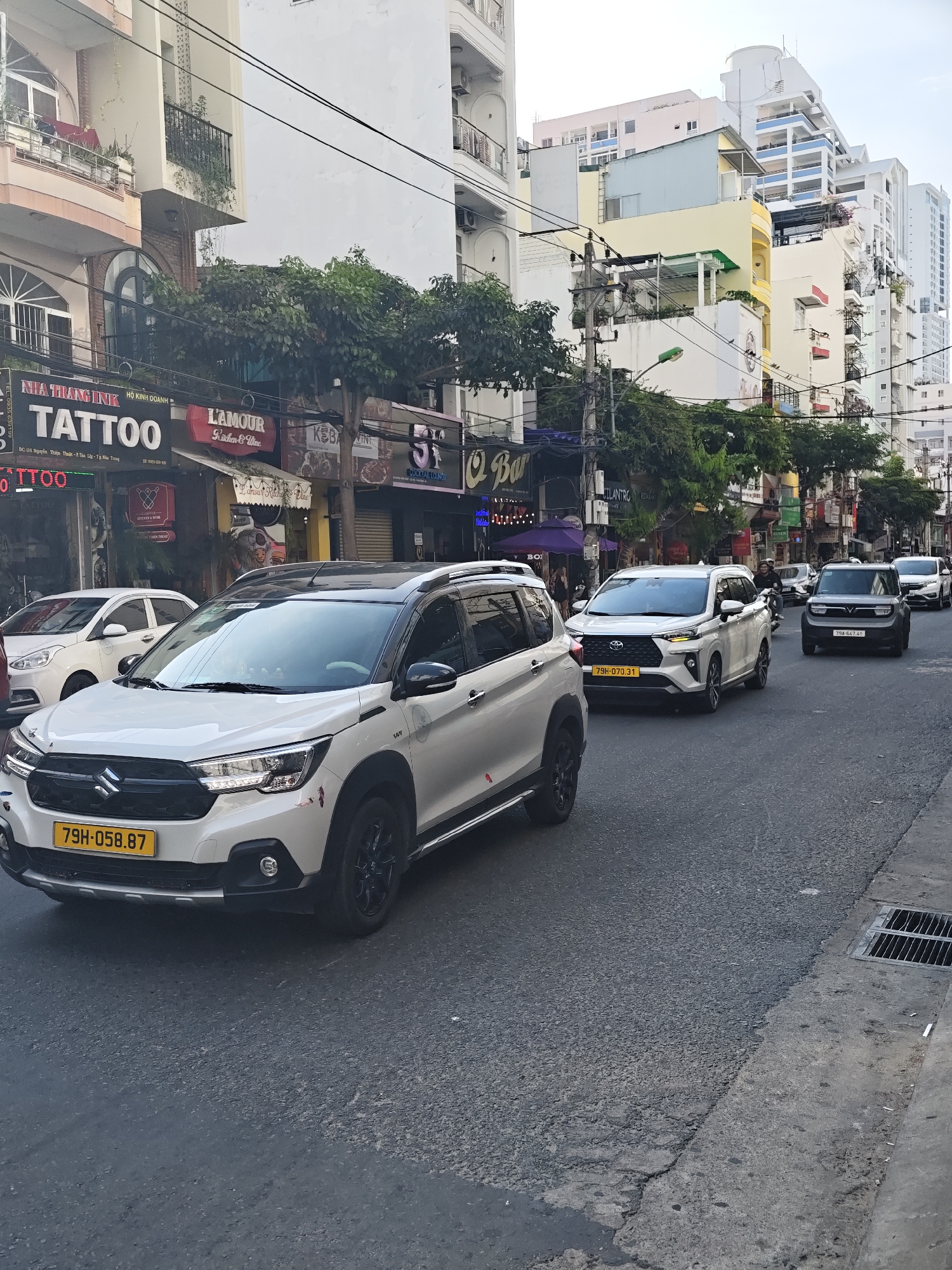 The image depicts a street scene in Nha Trang Ward, Vietnam, on a cloudy day. In the foreground, a white Suzuki X-Lander SUV with license plate 79H-058.87 is driving on the right side of the road. To its left, a white Toyota Vios is visible. Further down the street, a dark-colored VinFast Fadil SUV with license plate 79A-647.41 is also in traffic. A motorcycle is seen in the distance, also heading in the same direction.
The street is lined with multi-story buildings, many of which appear to be commercial establishments with signage indicating businesses such as "TATTOO," "L'AMOUR Kitchen & Wine," and "Q BAR." The buildings have balconies and are adorned with plants. Power lines and streetlights are visible overhead. The road surface appears to be asphalt with some cracks and patches. A metal grate for drainage is present on the right side of the road. The overall environment suggests a bustling urban area.