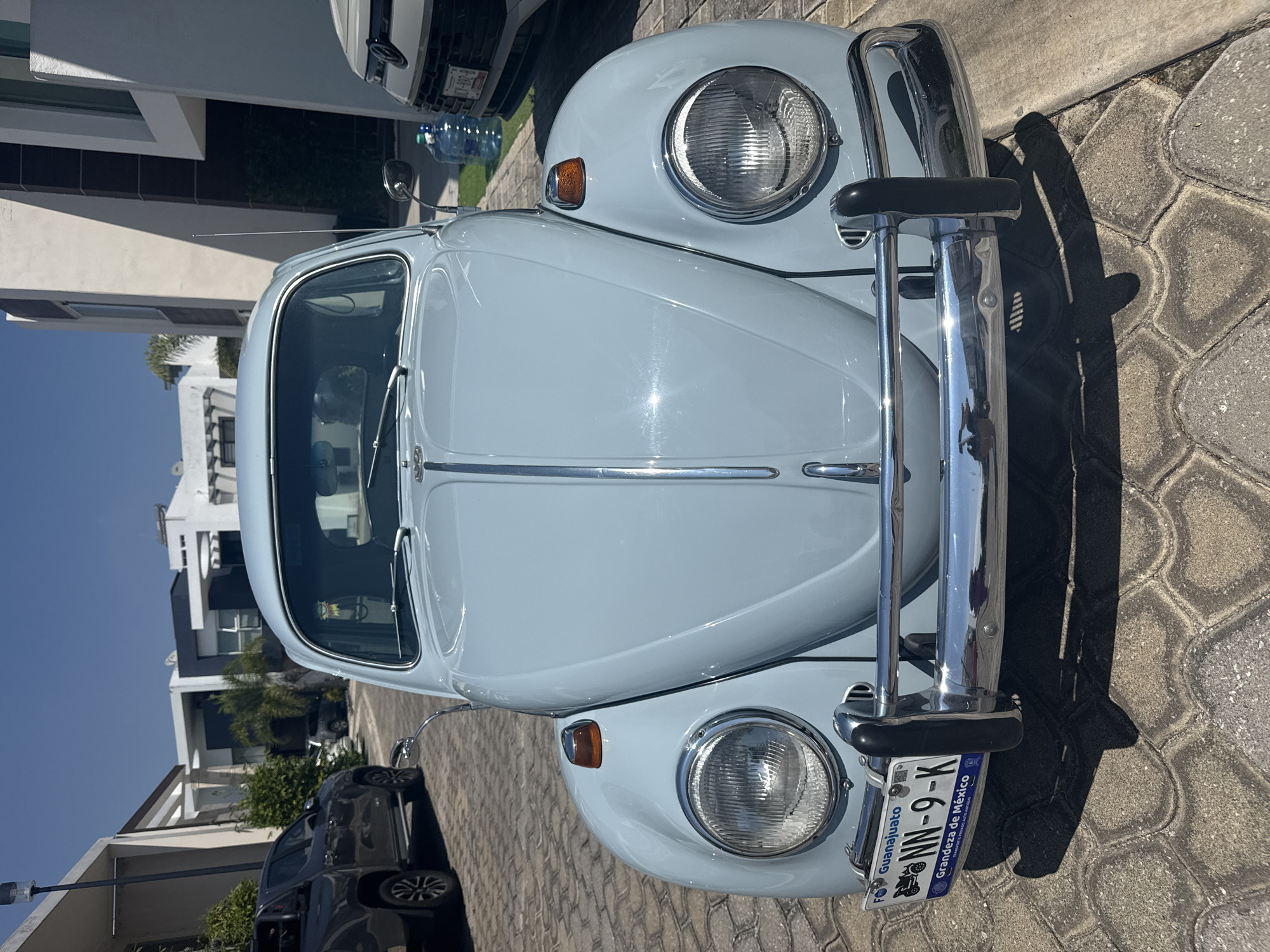 The image shows the front of a light blue Volkswagen Beetle parked on a paved surface, likely in Santa Clara Ocoyucan, Mexico. The car's license plate is visible, displaying "NN-9-K" and indicating the state of Guanajuato. The Beetle has chrome bumpers and headlights. In the background, modern houses and a dark SUV are visible, suggesting the car is parked on a residential street. The sky is clear and sunny. A white vehicle is parked directly behind the Beetle.