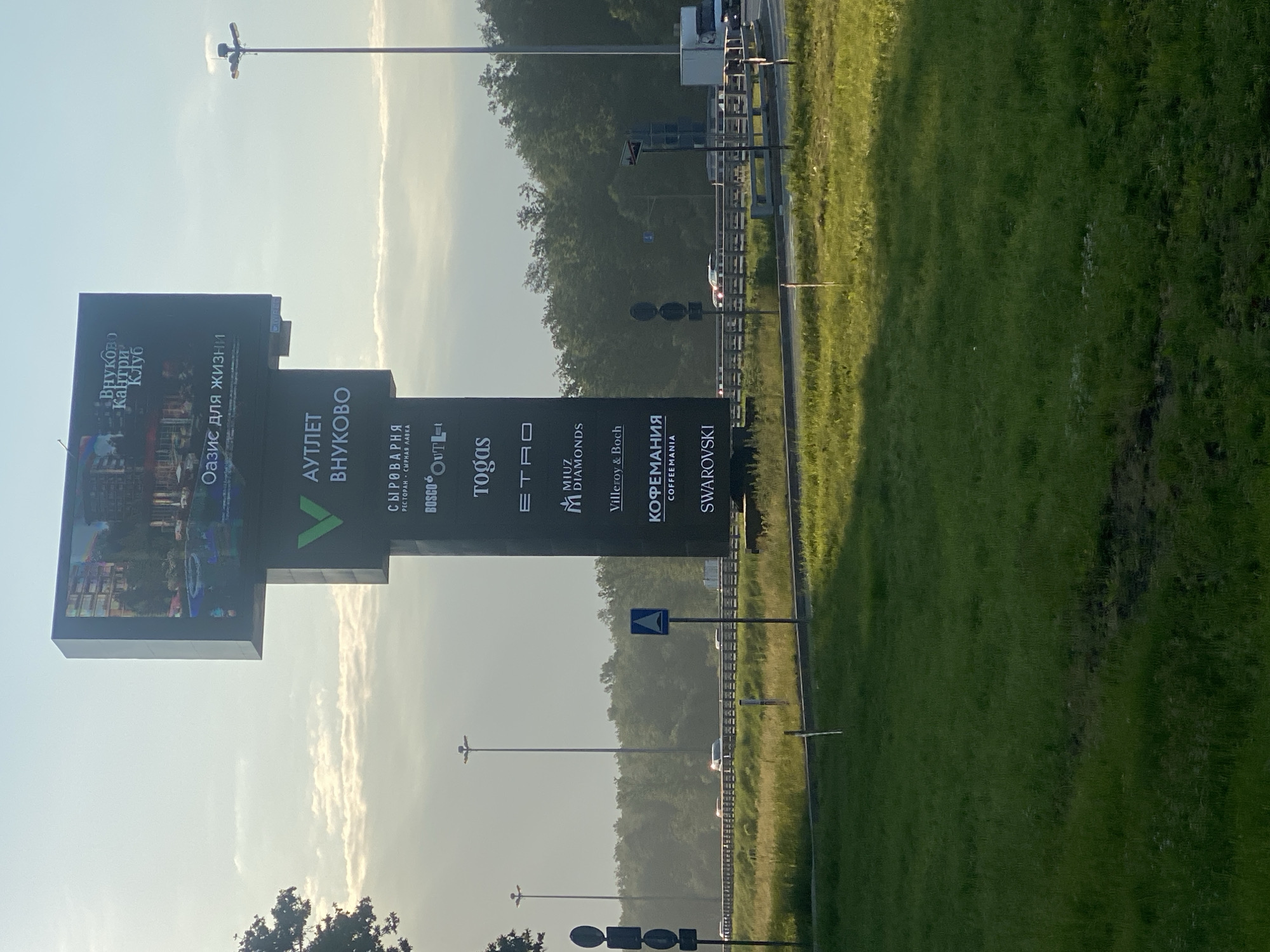 A tall, black billboard stands in a grassy area next to a multi-lane highway. The billboard features multiple sections with text and logos. The top section displays an image of a building with a pool and text in Russian. Below this, a large green "V" logo is visible, followed by more text in Russian. Further down, there are logos for various brands, including "BOSCO," "Togas," "ETRO," "MIUZ DIAMONDS," "Villeroy & Boch," "KOHEMANIA," and "SWAROVSKI." The highway has several vehicles on it, and trees line the side of the road in the background. The sky is overcast with some clouds.