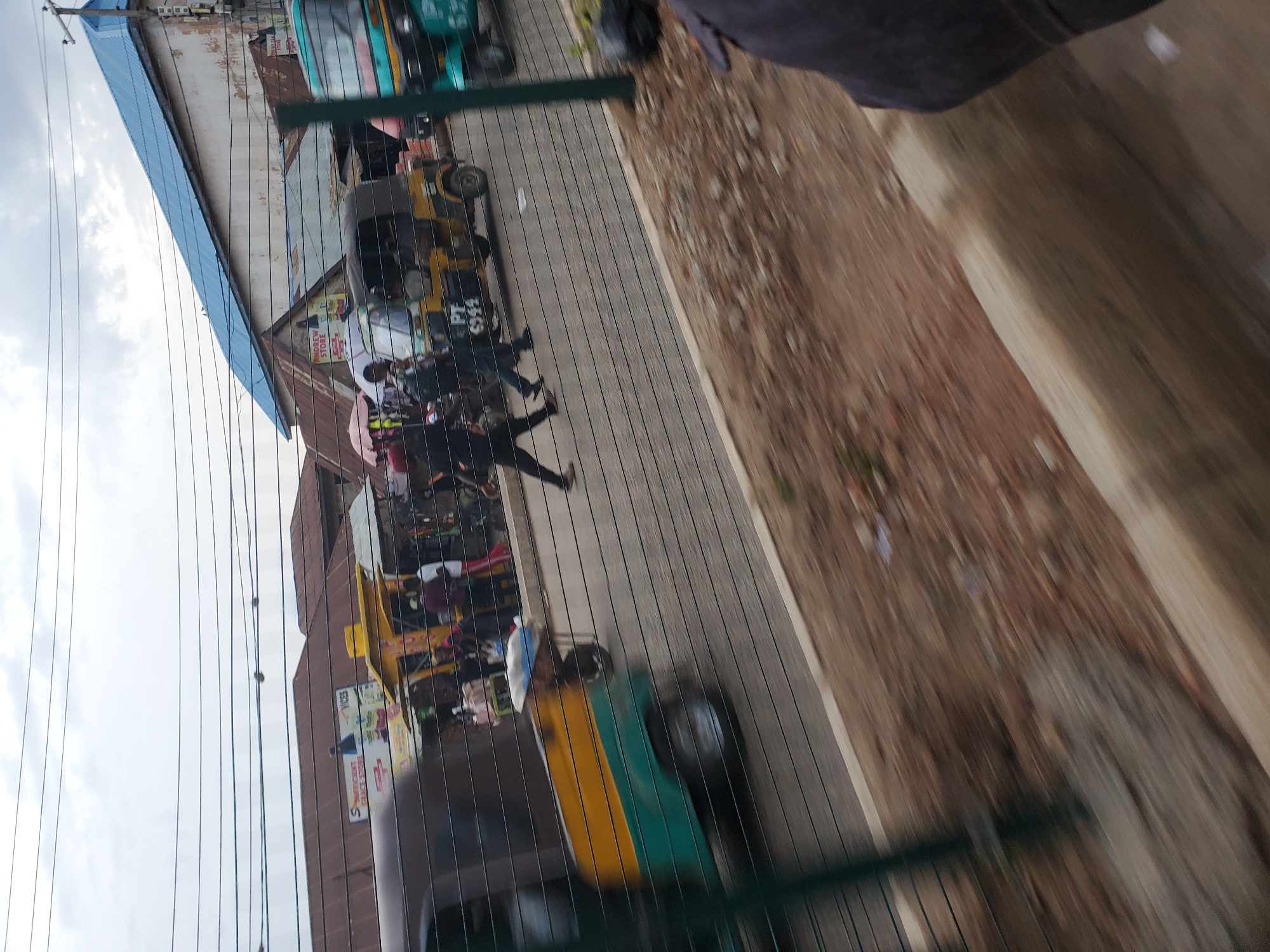 The image depicts a street scene in Bukuru, Nigeria, characterized by a bustling market atmosphere. Several motorized rickshaws, known as "Keke NAPEPs" in Nigeria, are visible, with one prominently featuring a yellow and green color scheme. Pedestrians are walking along the roadside, and some are interacting with vendors at market stalls. Buildings with corrugated iron roofs line the street, suggesting a commercial area. The sky is overcast, indicating a cloudy day. The overall impression is one of a typical urban marketplace in Nigeria, filled with activity and local transport.