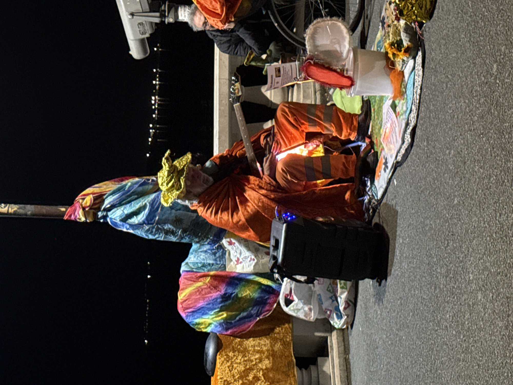 The image shows a man playing a guitar on the street at night. He is dressed in orange clothing and a golden headdress, sitting amongst colorful fabrics and other objects. Next to him is a black speaker. A person in dark clothing stands near a bicycle in the background. In the far background, faint lights suggest a cityscape. The image was taken in Burgas, Bulgaria.