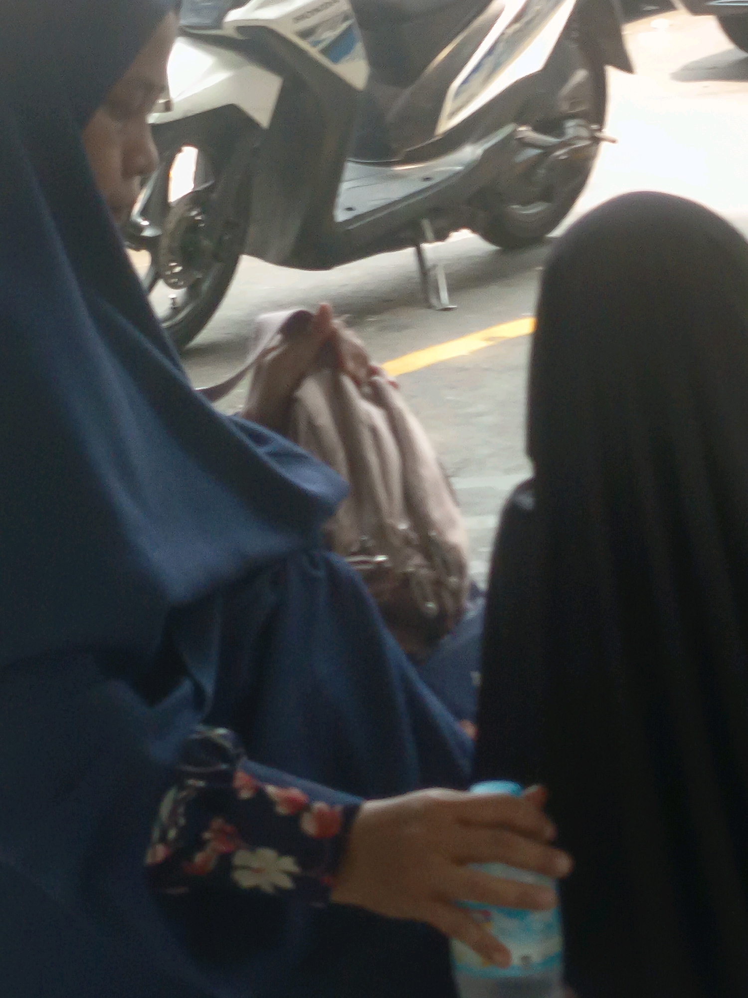 Two individuals, likely women based on their attire, are present. One, on the left, wears a dark blue hijab and a dark blue garment with a floral pattern on the sleeve. Her left hand is visible, holding a clear plastic bottle with a blue cap. The other individual, on the right, wears a black hijab and appears to be seated. In the background, a white and silver scooter is parked. The ground appears to be paved with a yellow line visible on the right side of the image. The setting suggests an outdoor urban environment.