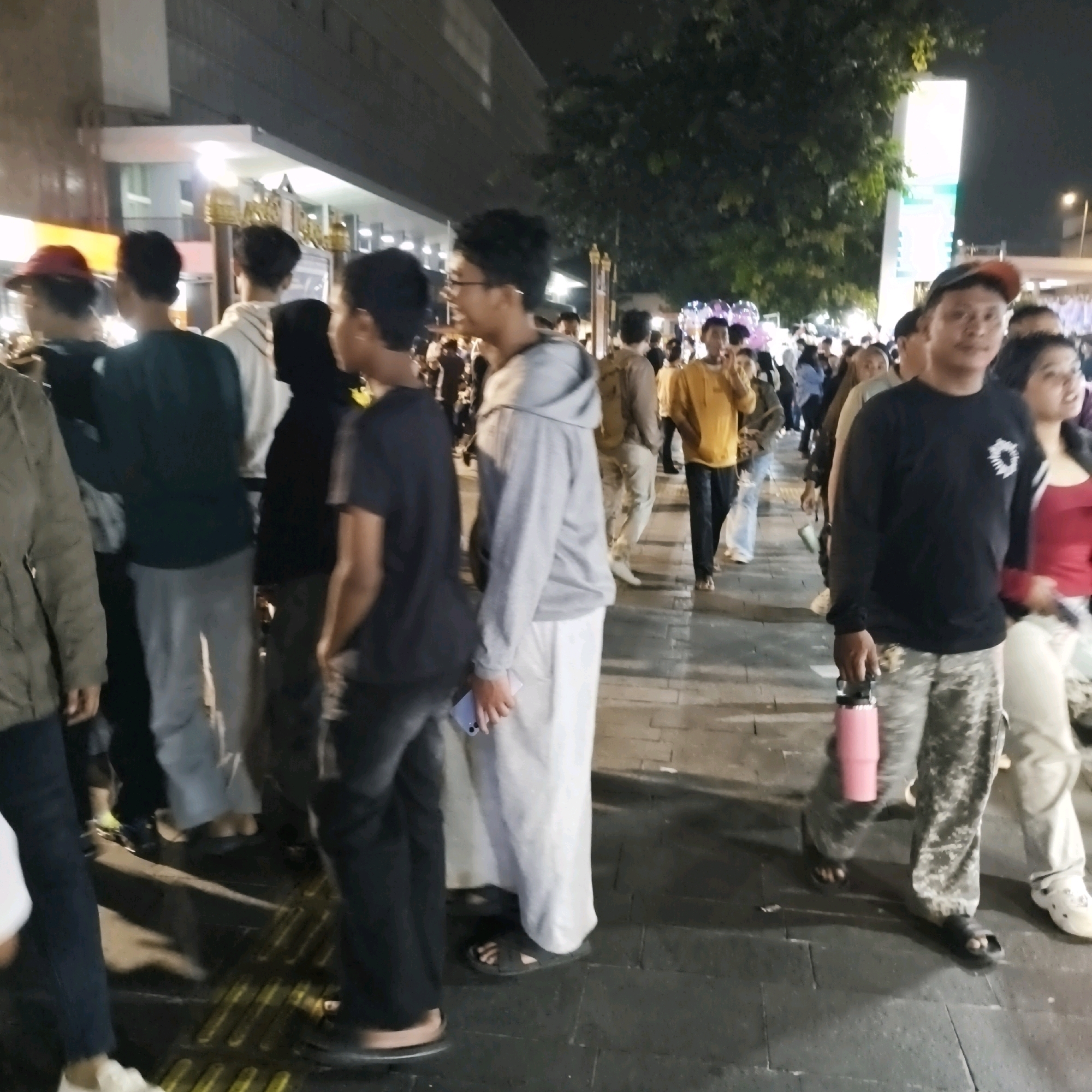 The image depicts a crowded street scene at night, likely in West Jakarta, Indonesia, given the context provided. A diverse group of people, predominantly young adults and teenagers, are walking on a tiled pedestrian walkway. They are dressed in casual, modern attire. The background shows building facades with illuminated windows, indicating commercial or residential spaces. Trees with foliage are visible, suggesting an urban setting with some greenery. Streetlights and the ambient glow from buildings illuminate the scene.
In the foreground, a person wearing a black long-sleeved shirt with a circular emblem and camouflage-patterned pants is holding a large pink water bottle. To their left, another individual in a grey hooded sweatshirt and white pants is seen from the side, holding a mobile phone. Other individuals are walking in various directions, forming a flow of pedestrian traffic. Some are looking ahead, while others are engaged in conversation. The ground appears wet, reflecting the artificial lights. The overall impression is one of a bustling urban environment at night, possibly during a public event or simply a busy thoroughfare.