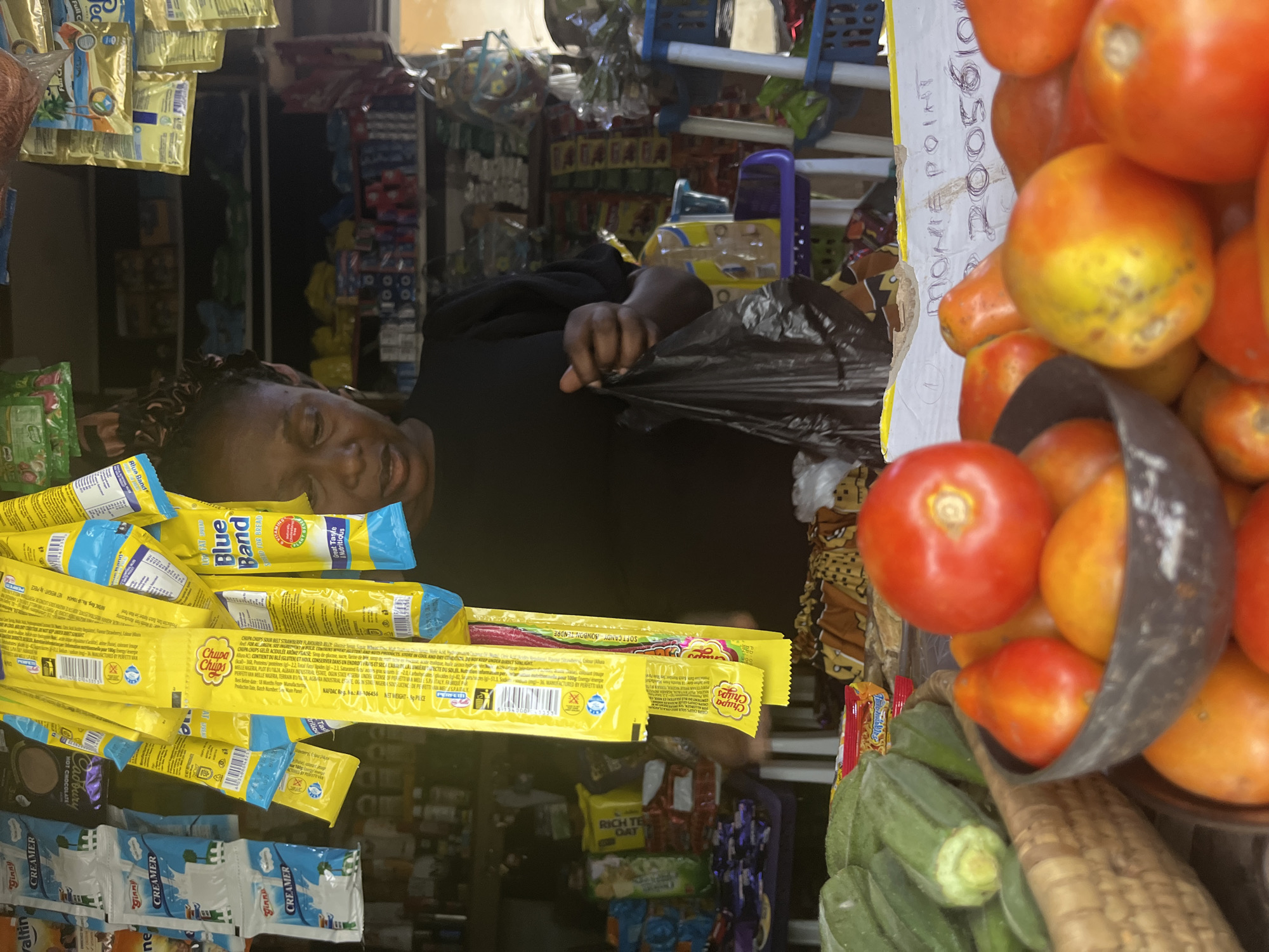 In a bustling outdoor market stall in Abuja, Nigeria, a woman with dark, braided hair wears a black top and patterned skirt. She is holding a black plastic bag in her right hand, her gaze directed downwards as if assessing goods. The stall is laden with an array of items: bright yellow packets of Blue Band spread and Chupa Chups lollipops hang prominently, interspersed with sachets of "Ginny" creamer and other packaged foods. Shelves behind her are stocked with various bottled and packaged products. In the foreground, a woven basket overflows with green okra, and a dark bowl contains ripe tomatoes and other red and orange fruits. A white piece of cardboard with handwritten text, including "MONIE POINT" and a series of numbers, is visible near the fruit. The scene captures a snapshot of commerce and daily life in the vibrant city.
