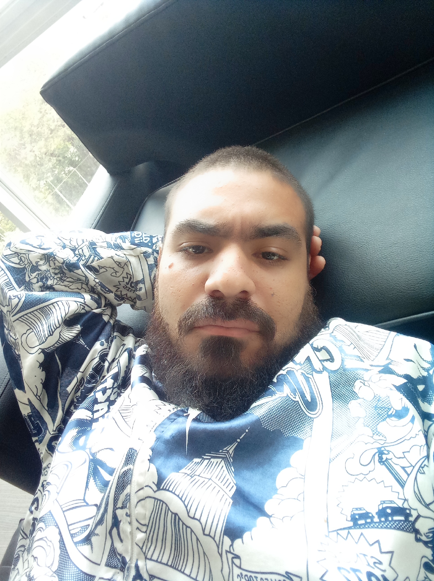 A man with short hair and a beard is reclining in a black chair. He is wearing a blue and white patterned shirt depicting cityscapes, possibly New York City, with buildings and cars. He has his hand behind his head. A window is visible behind the chair. The image appears to be taken indoors. The location in Parroquia Guatire, Venezuela, is not discernible from the image.