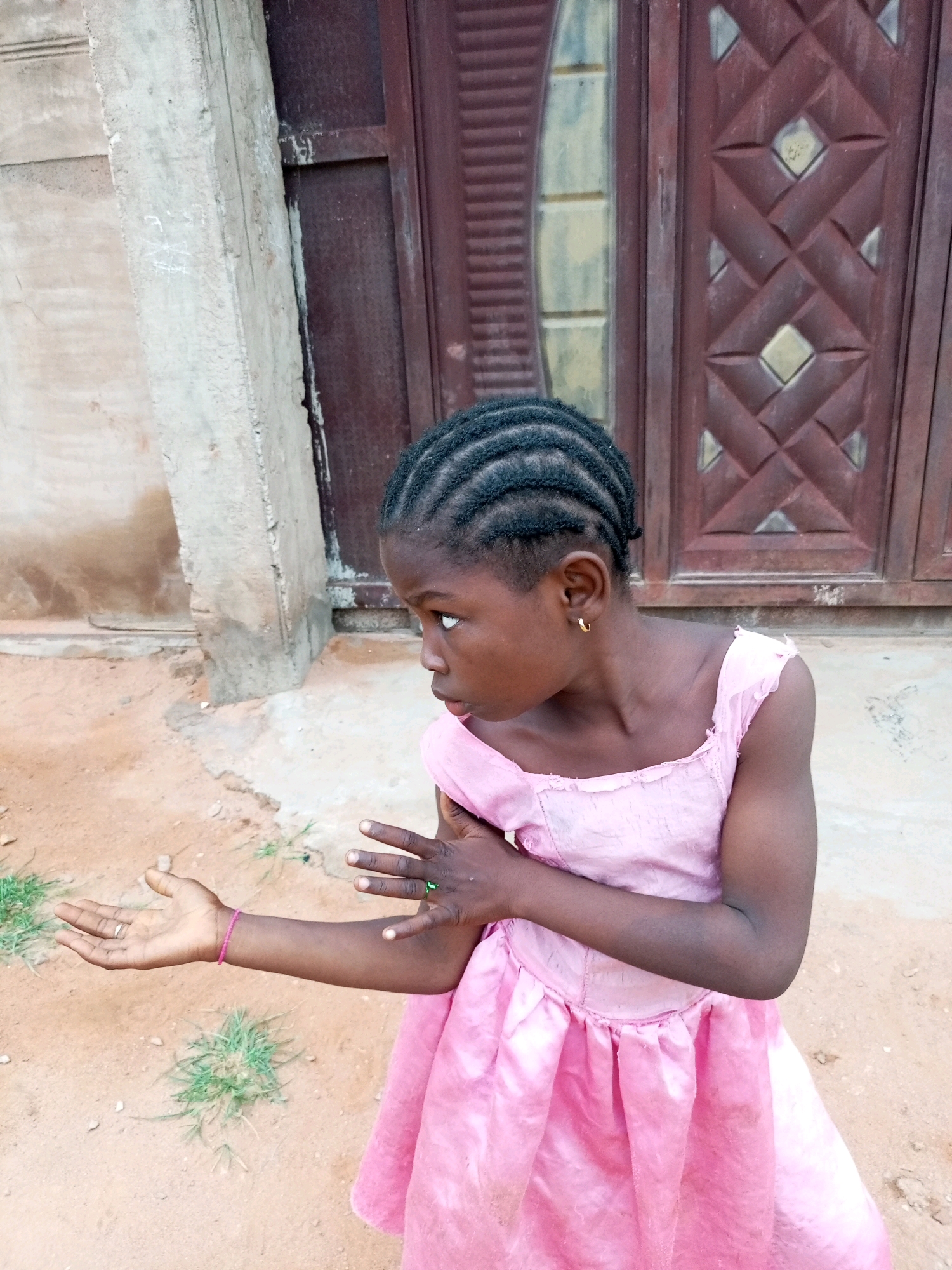 A young girl with braided hair and a pink dress stands outside on a dirt ground, facing left, in front of a brown wooden door. The girl's left arm is extended outwards, with her palm open. Her right arm is bent, with her hand on her chest. The environment appears to be a residential area in Tempure, Nigeria, with a concrete pillar to the left of the girl and some sparse vegetation on the ground.
