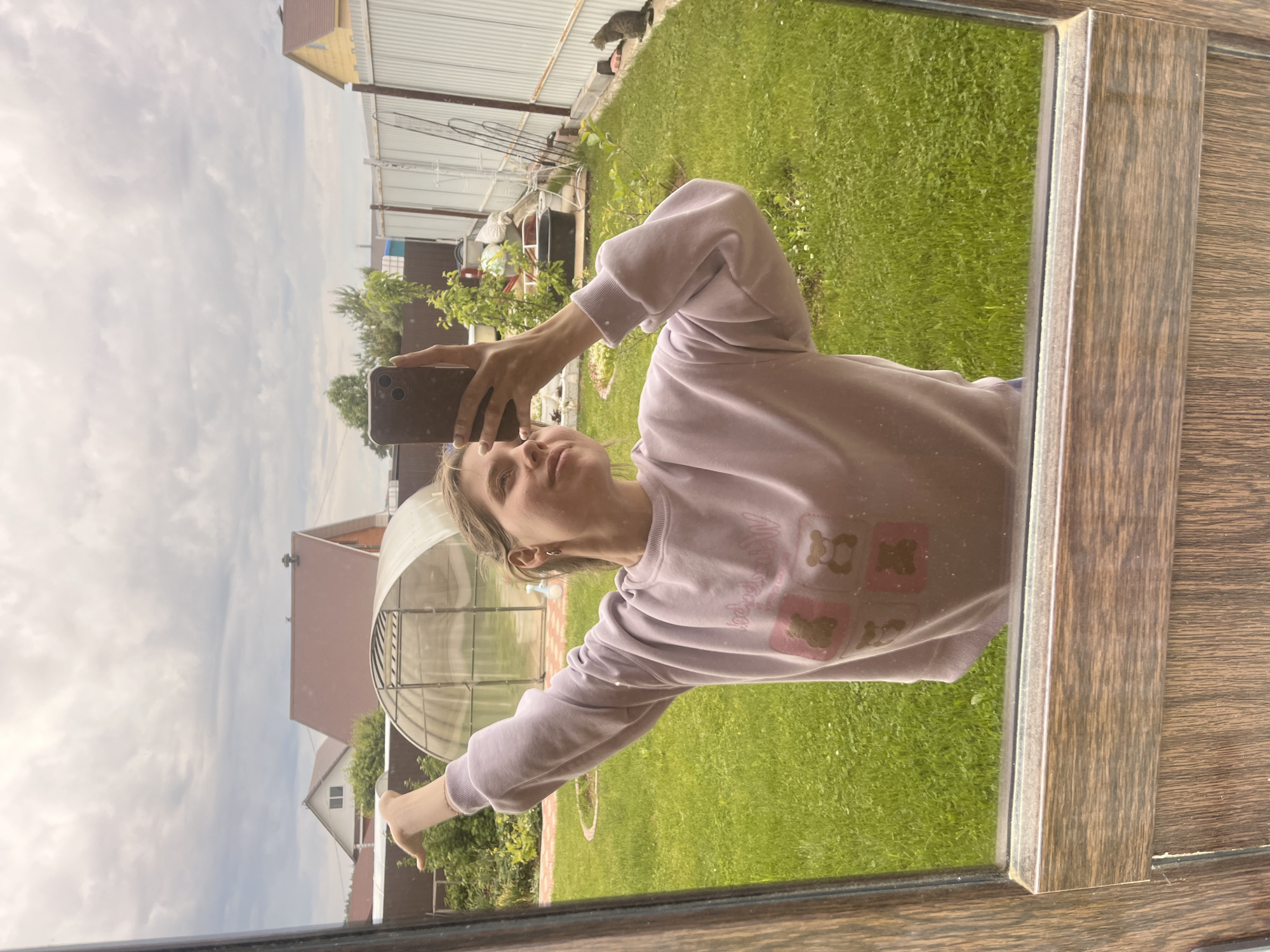 The image shows a person in a pale purple sweatshirt taking a selfie in a mirror. The mirror reflects a backyard scene, featuring a greenhouse, green grass, and a fence. The surrounding area includes houses and trees, suggesting a residential area. The exact location is reported as Chaikovsky, Russia.