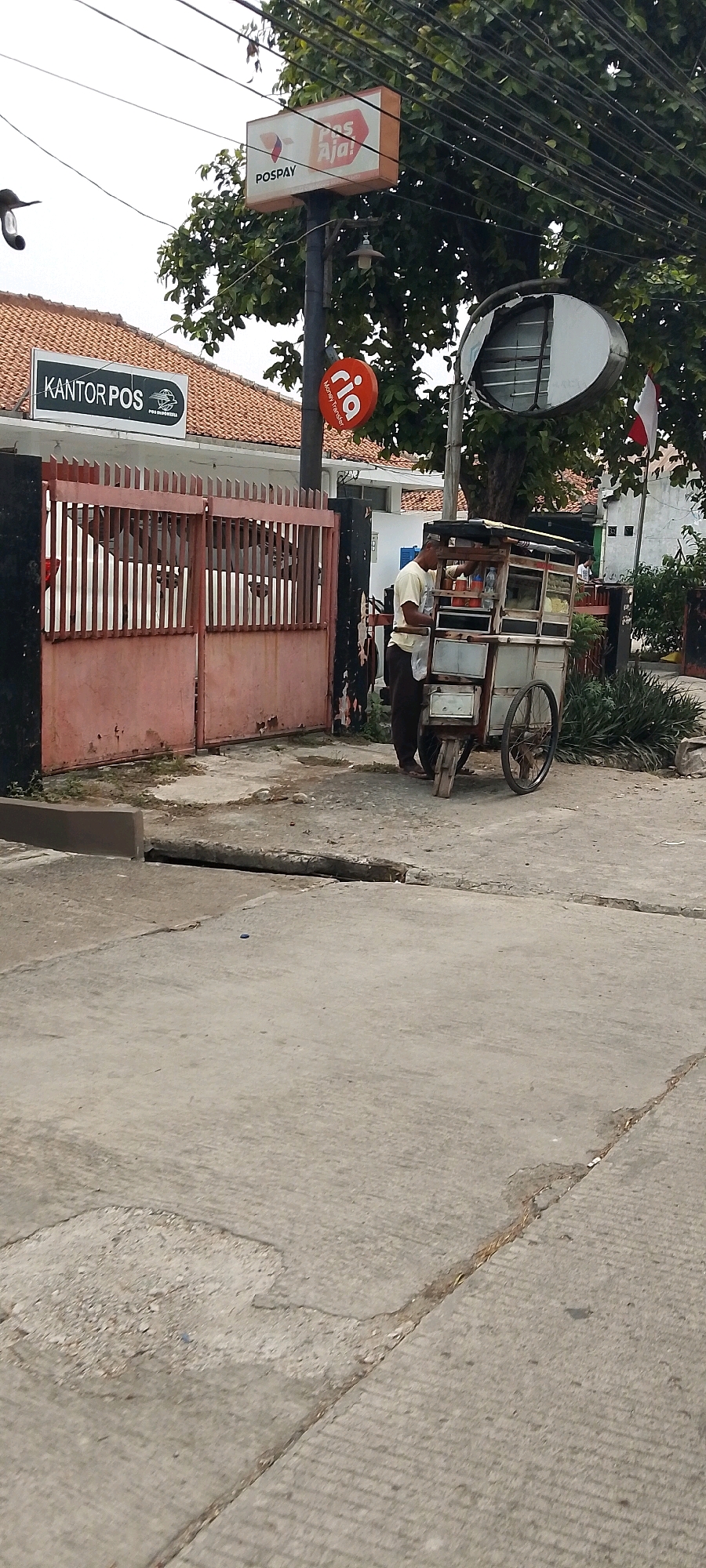 In Depok, Indonesia, a man is standing next to a food cart on a concrete street. The cart is made of wood and metal, with wheels and a canopy. The man appears to be interacting with the cart, possibly preparing or serving food. In the background, a building with a red-tiled roof and a sign that reads "KANTOR POS" (Post Office) is visible. Another sign above the post office indicates "POSPAY". A circular red sign with the word "ria" and "Money Transfer" is also present. The scene is set outdoors, with trees and electrical wires overhead. The overall environment suggests a commercial or public area.