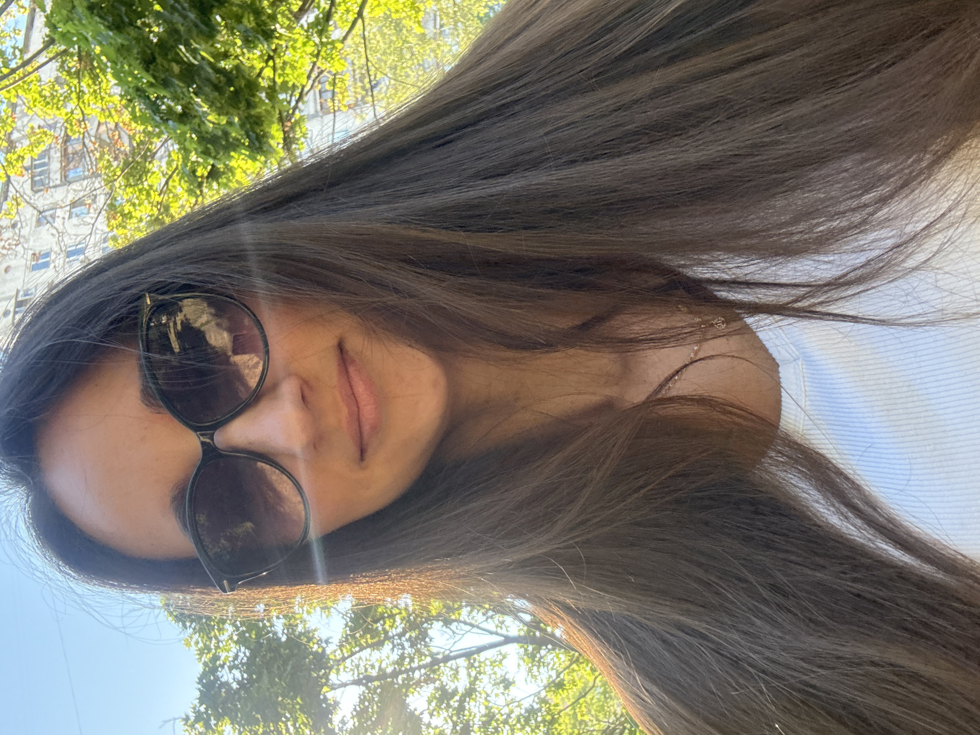 In a frontal, head-and-shoulders shot, a woman with long, dark brown hair and fair skin is captured outdoors, likely in an urban environment. She is wearing large, dark sunglasses, obscuring her eyes, and a white, ribbed top. A delicate gold necklace with a small pendant is visible around her neck. The background features bright green foliage from trees, illuminated by sunlight, and a light-colored, multi-story building with visible windows. The sky is clear and blue. The overall impression is of a sunny day in a city like Kyiv, Ukraine, given the architectural style of the building and the lush greenery.