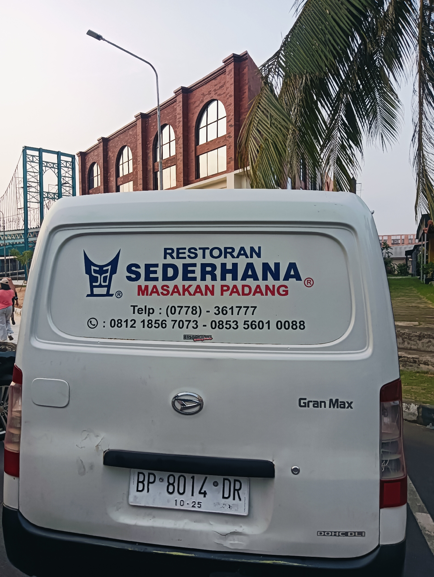 A white van is parked on a street. The van has the words "RESTORAN SEDERHANA MASAKAN PADANG" and a phone number printed on its back. A license plate with the letters "BP" and numbers "8014" is visible below the text. The van's rear bumper is black, and the surrounding area appears to be a road with a sidewalk and some greenery. In the background, a brick building with arched windows and a palm tree are visible. A lamppost is also present in the background.