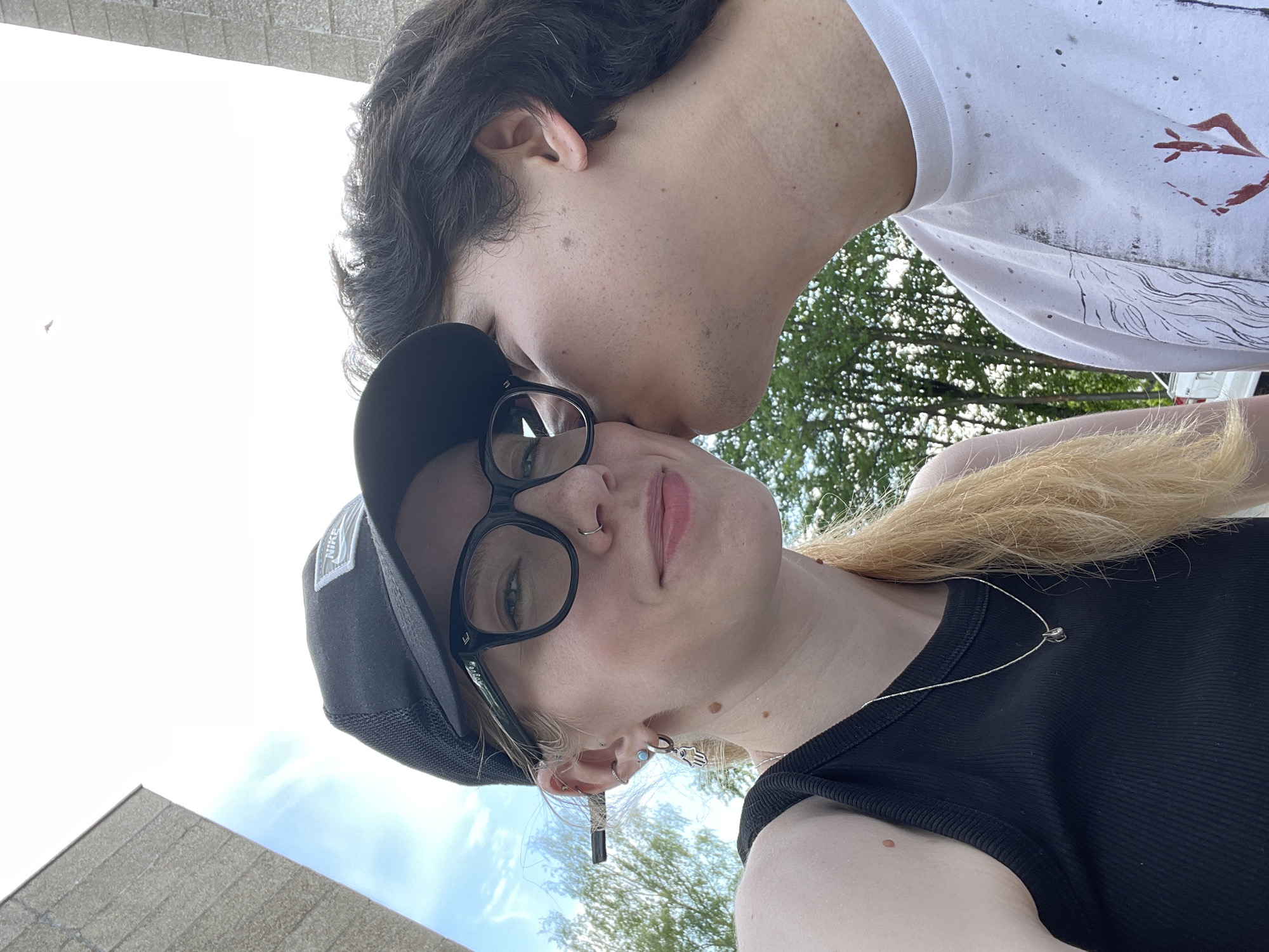 A selfie shows a young woman wearing a black tank top, a black Nike baseball cap, and black glasses with a nose ring and several earrings, one of which is a Hamsa hand design. She has blonde hair. A man is kissing her cheek. He is wearing a white t-shirt with a design and has dark hair. Trees and a building are visible in the background, suggesting an outdoor location. There is no visual information in this image to confidently conclude that the location is in the city of Izhevsk, Russia.