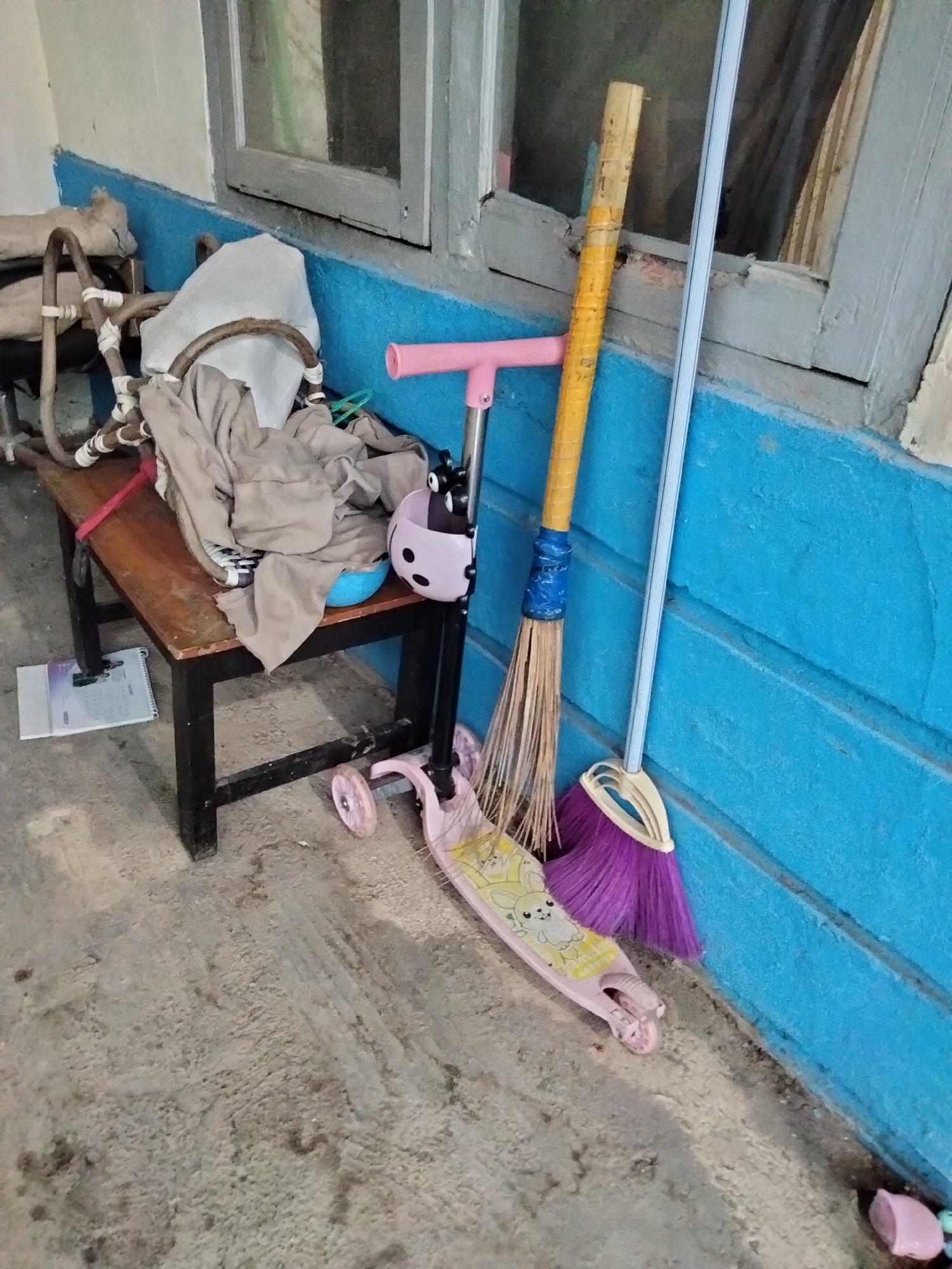 A pink children's scooter and a broom are leaning against a bright blue wall adjacent to a wooden bench. The scooter is decorated with a cartoon bunny. The broom has a yellow wooden handle and purple bristles. A pile of beige fabric, possibly blankets or clothes, is draped over the bench. To the left of the bench, a metal structure, possibly part of a baby stroller or chair, is visible. A notebook with a spiral binding lies on the ground in front of the bench. The scene is outdoors, with a concrete floor and a window above the blue wall. The overall impression is one of outdoor storage or a disused area.