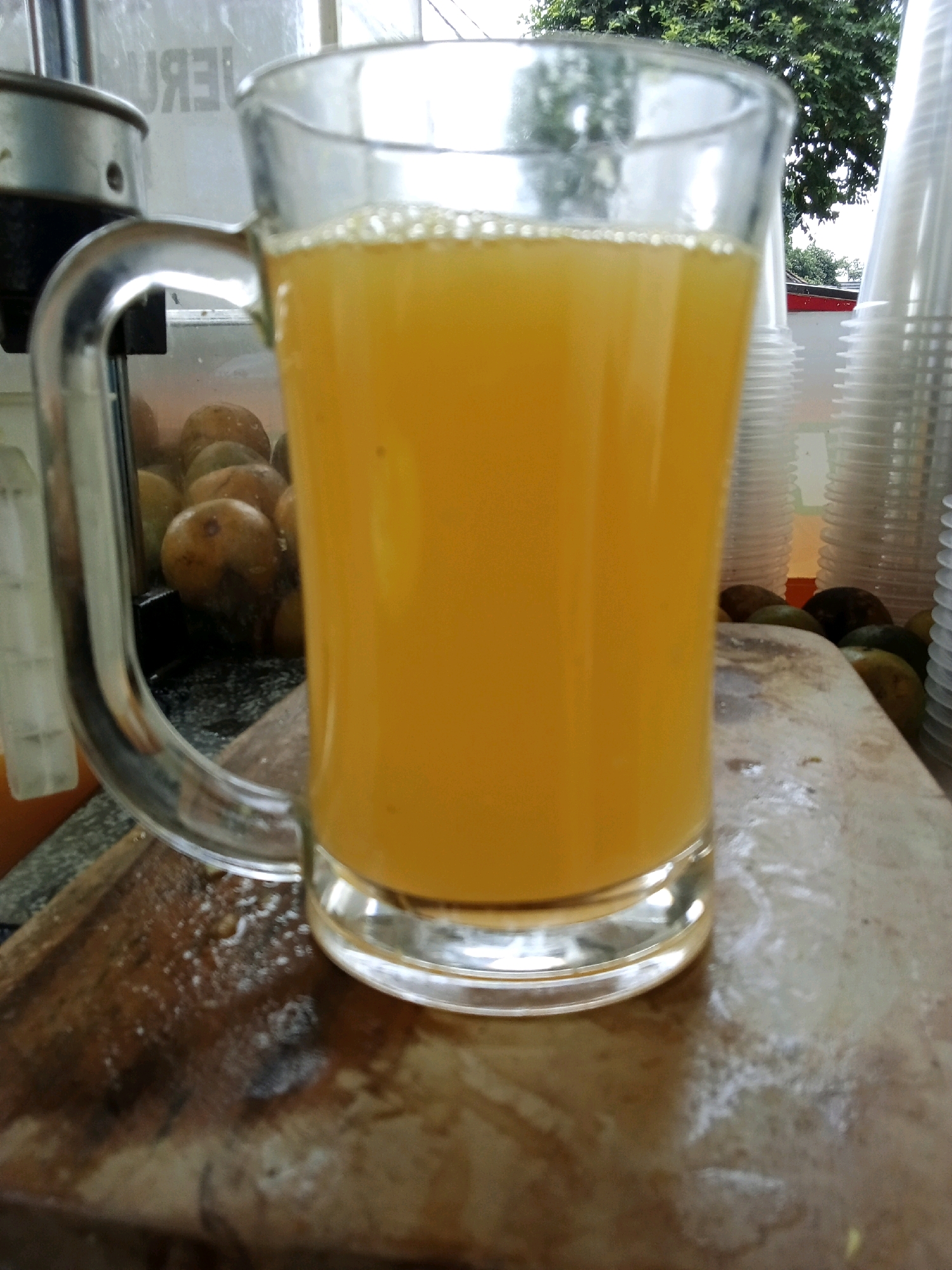 The image shows a close-up shot of a glass filled with orange juice, placed on a wooden surface. The glass has a handle and is filled to the brim, with a layer of foam on top. In the background, there are several oranges visible, suggesting that the juice may be freshly squeezed. There are also stacks of white plastic cups and trees in the background. The lighting suggests it is daytime, and the environment appears to be an outdoor stall or shop. While the image itself does not provide specific details about its location in South Jakarta, Indonesia, this type of scene is commonly found in street food stalls or juice vendors throughout the city, especially in areas known for their culinary offerings.