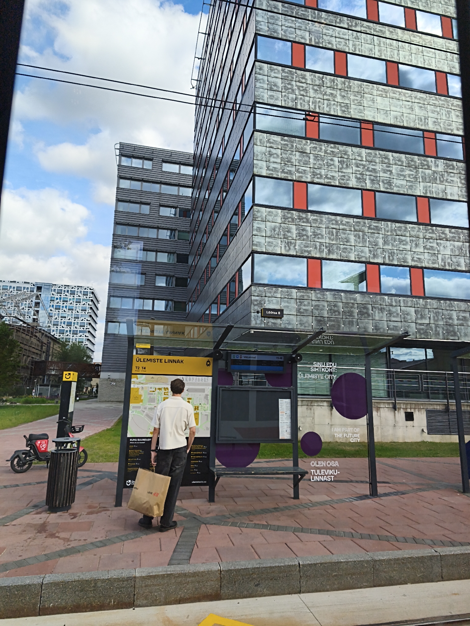 The image depicts a street scene in Tallinn, Estonia, specifically at the Ülemiste City business district. In the foreground, a man wearing a light-colored polo shirt and dark jeans stands at a bus stop, holding a brown paper shopping bag from H&M. He is facing a large information board with a map of the area and public transport information. To his left, a red electric scooter is parked next to a trash can. Behind the bus stop shelter, a modern, grey building with red accents and numerous windows reflects the sky. Further in the background, another contemporary building with a grid-like facade is visible. The ground is paved with reddish-brown bricks and grey stone. The sky is partly cloudy.
