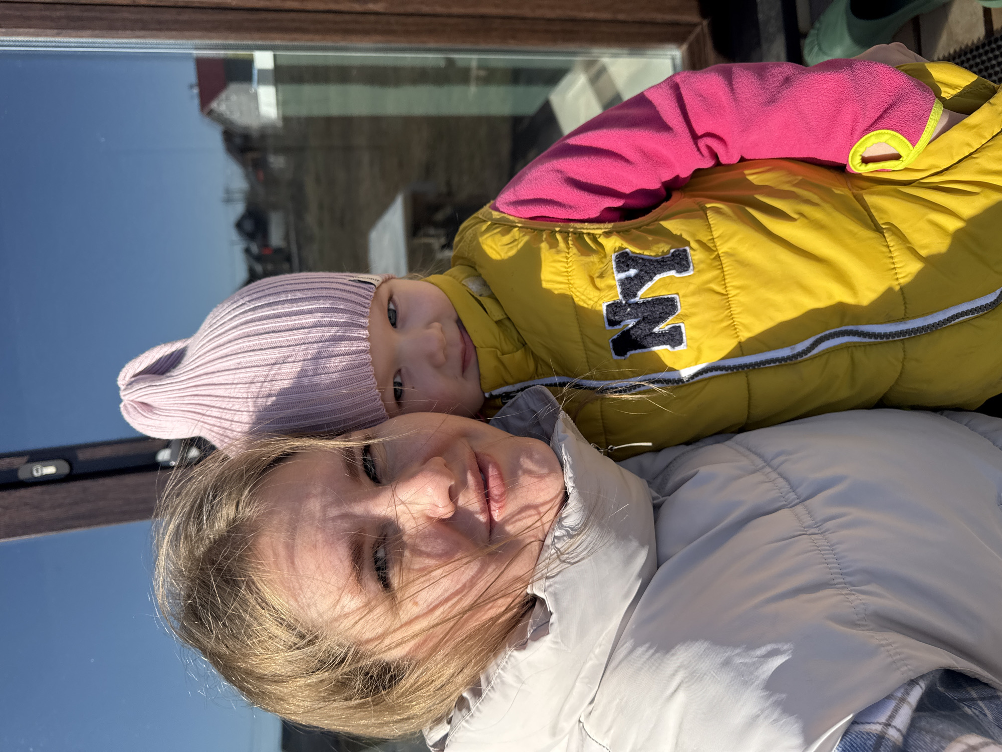 The image shows a woman and a baby taking a selfie. The woman has blonde hair and is wearing a white jacket. The baby is wearing a pink hat, a yellow jacket with the letters "NY" on it, and a pink undershirt. They appear to be outside, near a window reflecting a building and the sky. The picture was taken in Gornyi, Dvurechenskoe S/P, Russia.