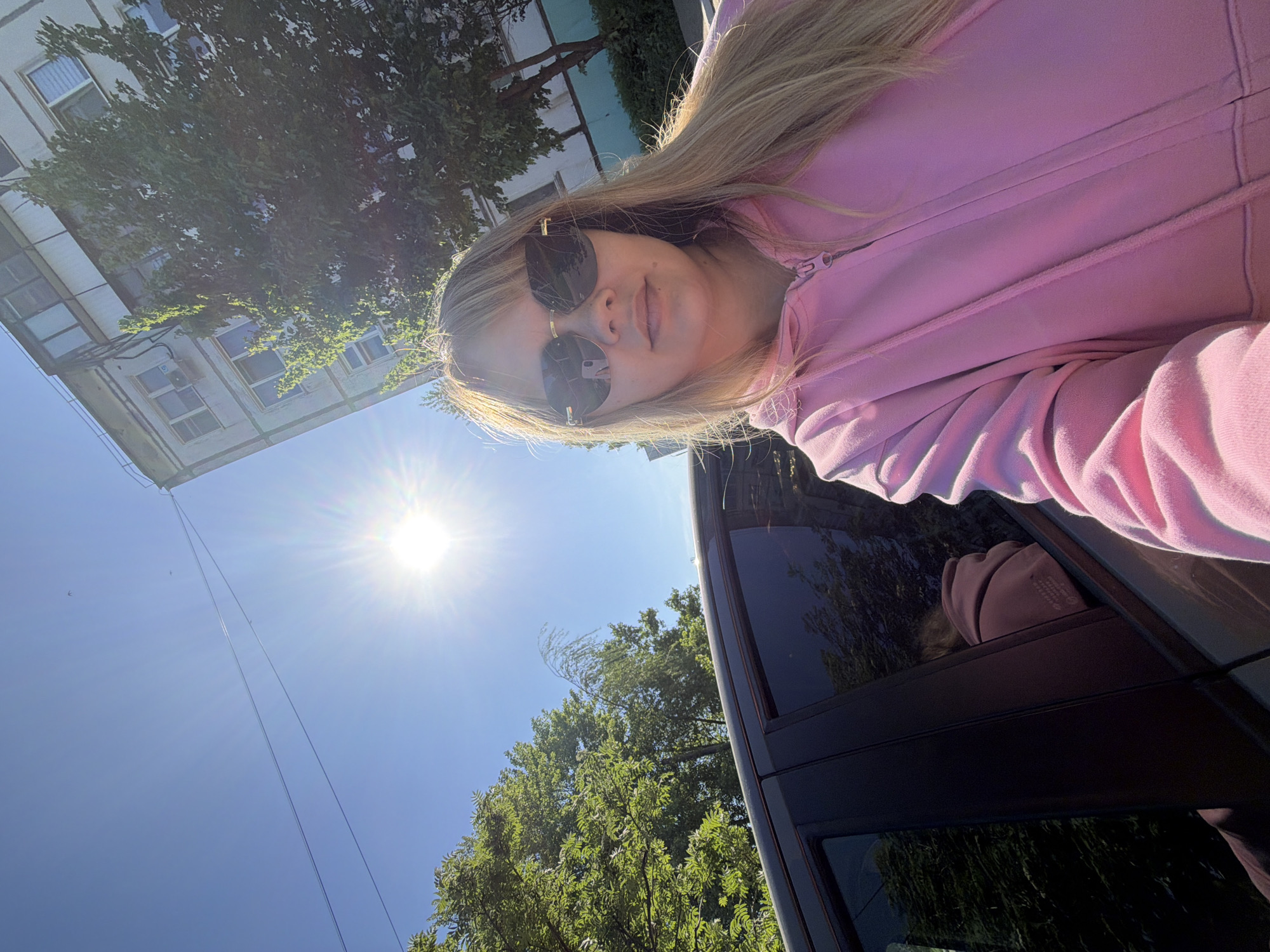 A low-angle, outdoor selfie shows a woman with blonde hair and sunglasses wearing a pink hooded sweatshirt. The sun is visible in the sky. Behind her are trees and part of a building with multiple windows. Part of a vehicle is visible to the right. The location appears to be the city of Chaikovsky, Russia.