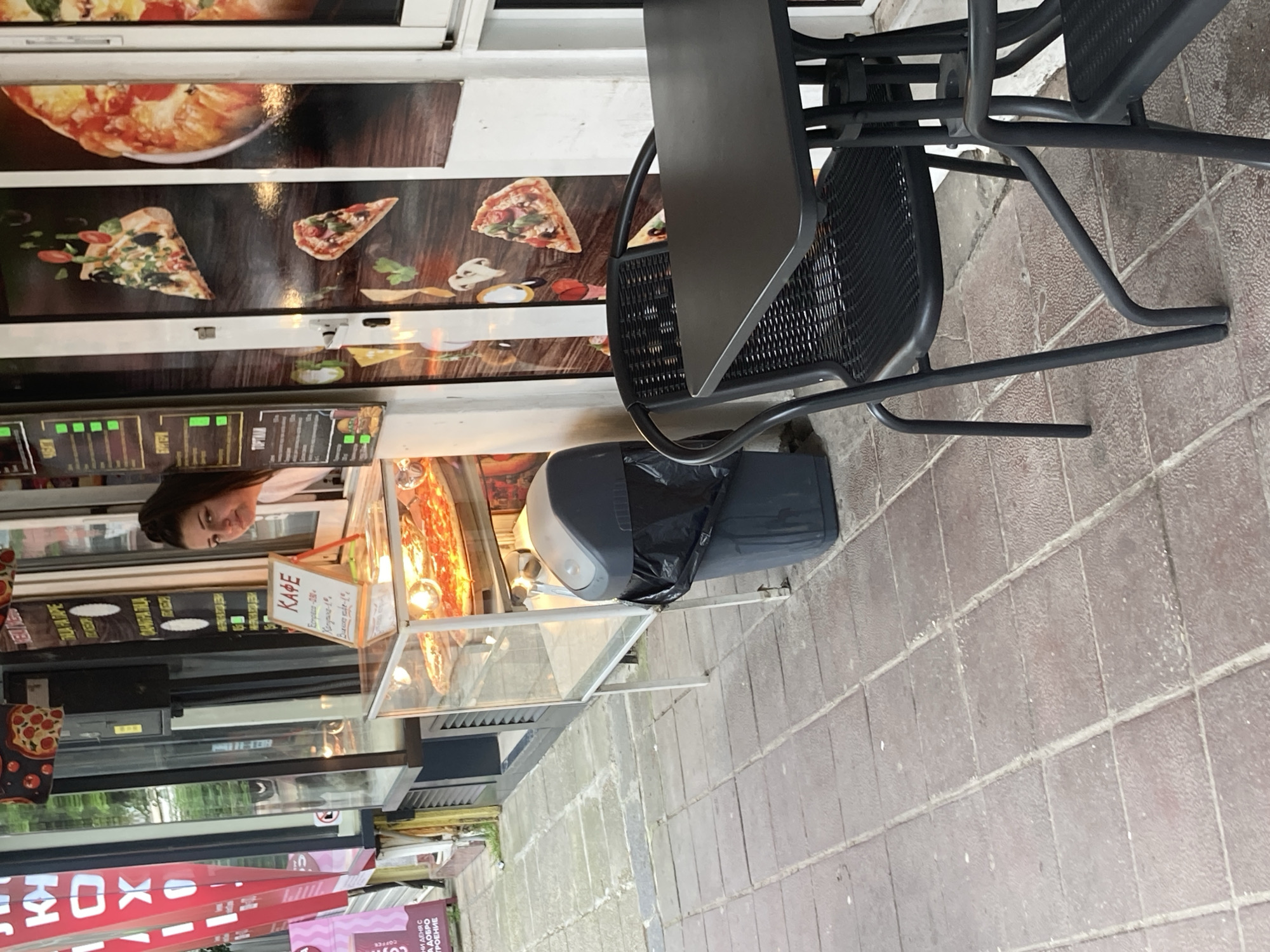 Here is a forensic description of the image:
**Location:** The image appears to be taken on a city street in Varna, Bulgaria. Evidence for this comes from the Cyrillic text "ΚΑΦΕ" and "ХОХ" signs, common in Bulgaria.
**Scene Overview:** The photo shows a street-level view of a small outdoor eatery or pizzeria. A black metal table and chairs are prominent in the foreground. Behind them is a storefront with pizza images displayed on the windows. A glass display case shows food items and pricing on the counter.
**People:** A female individual is visible, likely an employee.
**Objects:**
* Pizza-themed window decals
* Metal table and chairs
* Glass display case with food
* Pricing signage
* Rubbish Bin
* Advertising material for coffee
**Environment:** The scene takes place on a paved sidewalk. The building seems to have a storefront with a glass display, suggesting the sale of food items.