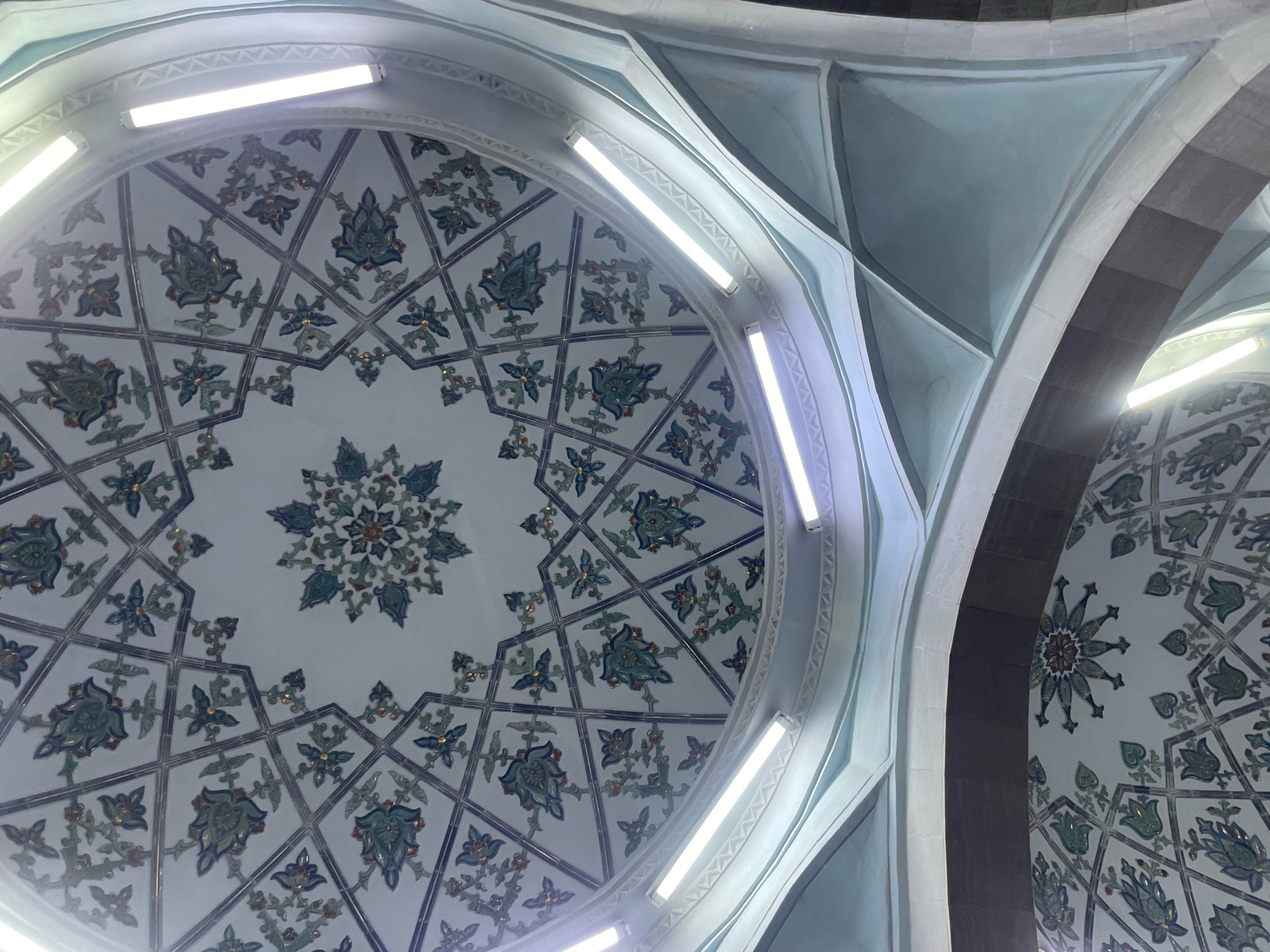The image shows the interior of a domed ceiling, likely in a mosque or historical building in Tashkent, Uzbekistan. The dome is decorated with intricate geometric patterns and floral motifs in shades of blue, green, and gold against a white background. Several fluorescent light fixtures are attached to the ceiling, illuminating the detailed artwork. The architectural style features curved lines and arches, contributing to the grandeur of the space. No people or specific actions are depicted in the scene.