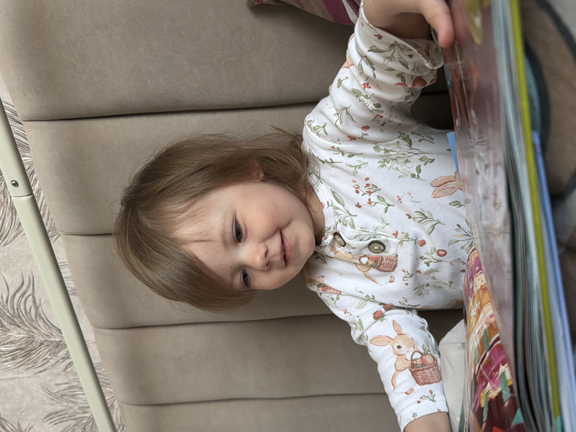 A young child is lying on a beige sofa with a book. The child is wearing a white long-sleeved shirt with a floral and rabbit pattern. The child is looking at the book and smiling. The sofa is upholstered in a tufted beige fabric. There is a white metal rod in the left side of the image. The location is assumed to be Reshetnikovo, Russia.