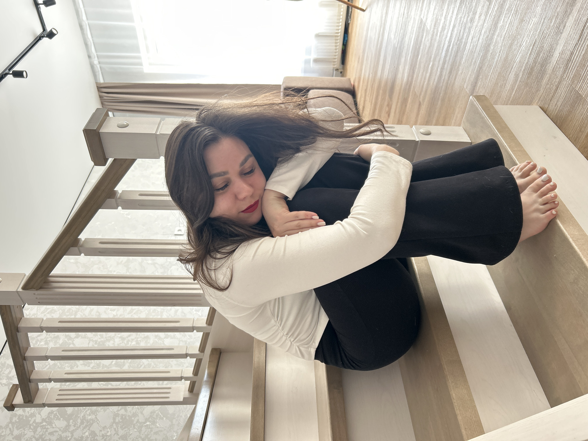 This image, taken in Chaikovsky, Russia, features a young woman sitting on a wooden staircase in an indoor setting. The image is rotated 90 degrees counter-clockwise from its natural orientation.
The subject is a woman with long, dark hair, light skin, and red lipstick. She is dressed in a plain white long-sleeved top and black wide-leg pants. She is seated with her knees drawn up towards her chest, her arms wrapped around her legs, and her head resting on her arms. Her eyes are cast downwards, suggesting a moment of quiet introspection, contemplation, or relaxation. Her toenails are painted white.
The scene takes place on a modern wooden staircase, characterized by light wooden treads and white risers. The banister on the left side of the frame also features light wooden elements. In the background, there is a large window letting in ample natural light, partially covered by light beige curtains. A white wall is visible behind the staircase, along with a sleek black track light fixture with multiple spotlights. Further into the room, a wooden floor with visible planks can be seen, and a radiator is partially visible beneath the window.
The bright natural light indicates it is daytime. The overall ambiance is calm and private, with a clean and minimalist interior design. There is no visible text in the image.