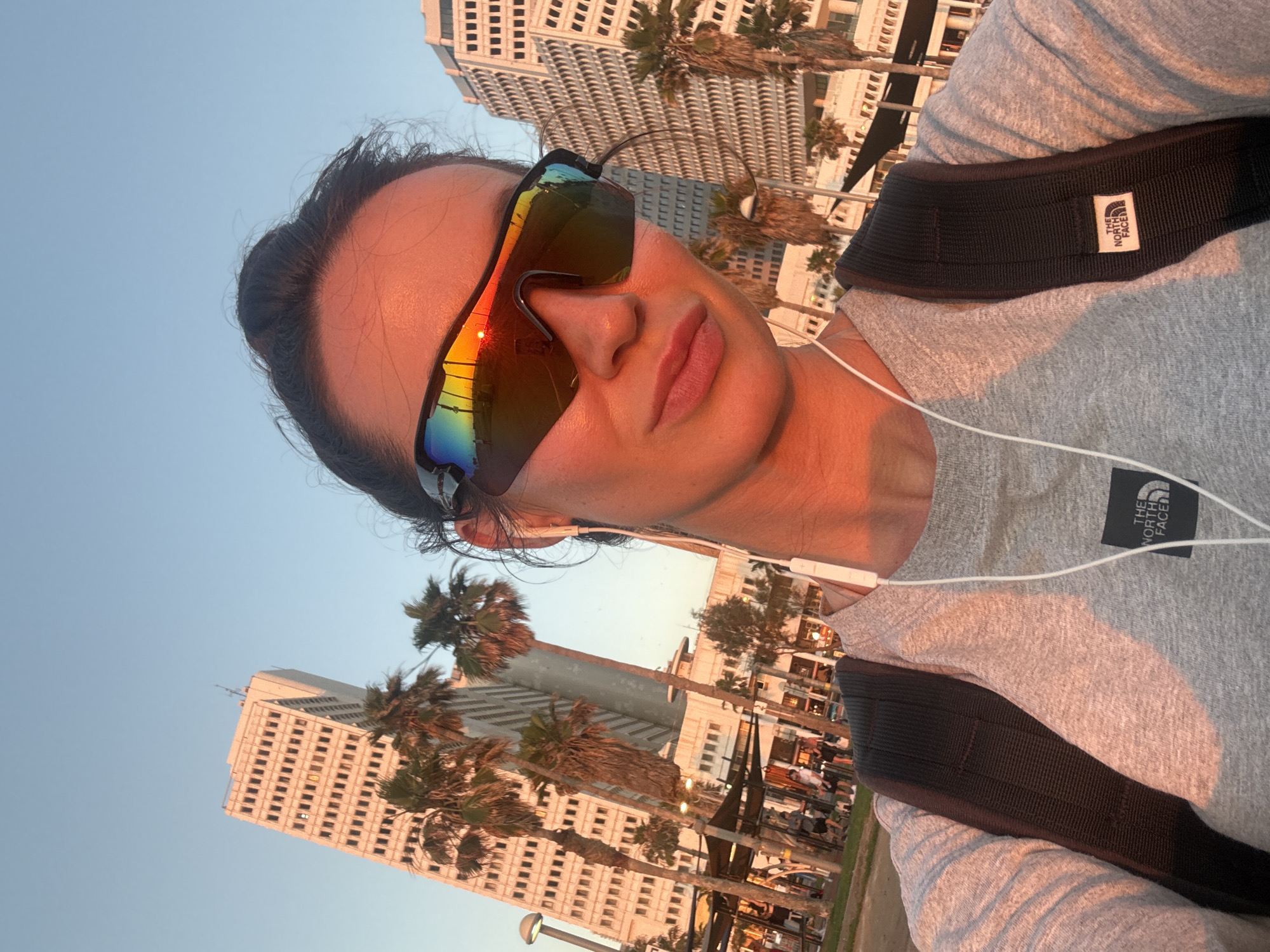 The media file shows a selfie taken outdoors, with the subject's face and upper torso prominently featured. The subject is wearing athletic sunglasses with a rainbow-colored lens and a grey t-shirt. White earbuds are visible, suggesting the subject is listening to music or a podcast. A black backpack strap with a "THE NORTH FACE" logo is also visible. The image is taken during what appears to be sunset, with warm, golden light illuminating the subject's face and casting long shadows. The background features tall buildings and palm trees, consistent with an urban environment in a warm climate. The scene suggests the subject is likely engaged in an outdoor activity, possibly running or walking, given the athletic attire and backpack. The overall mood is relaxed and candid, with the warm lighting contributing to a pleasant atmosphere. The text "THE NORTH FACE" is visible on the backpack strap.