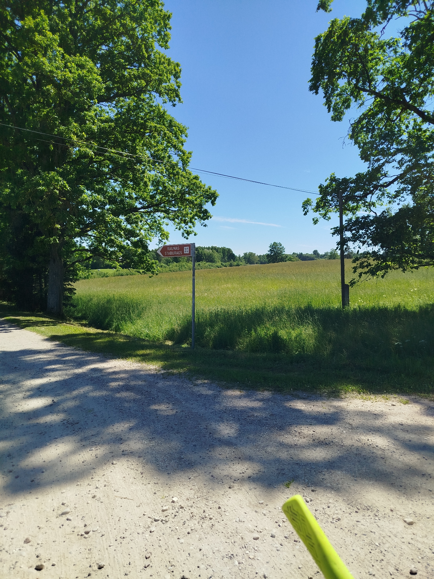 A dirt road with gravel and sand leads into a sun-dappled area, bordered by lush green trees on the left and right. The road is partially shaded by the trees, creating a pattern of light and shadow. In the middle ground, a tall signpost points to the right, indicating "RAUNAS STABURAGS" and a small graphic of a building. Beyond the sign, a vast field of tall green grass stretches towards the horizon, dotted with a few scattered trees in the distance. The sky above is a clear, bright blue. A bright yellow object, possibly part of a tool or equipment, is visible in the lower right corner of the image, partially obscuring the road.