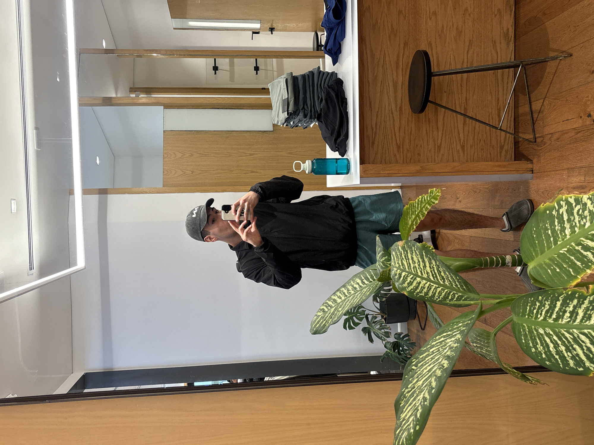 A man is taking a mirror selfie in what appears to be a retail store dressing room. The man is wearing a black jacket, green shorts, gray shoes, and a baseball cap. He is holding a phone to take the picture. Reflected in the mirror are shelves with folded clothing and a water bottle. To the right is a small stool and a plant with variegated green and white leaves. The environment suggests a modern and well-lit store interior. Without additional information, I cannot determine whether this photo was taken in the city of Puebla, Mexico.