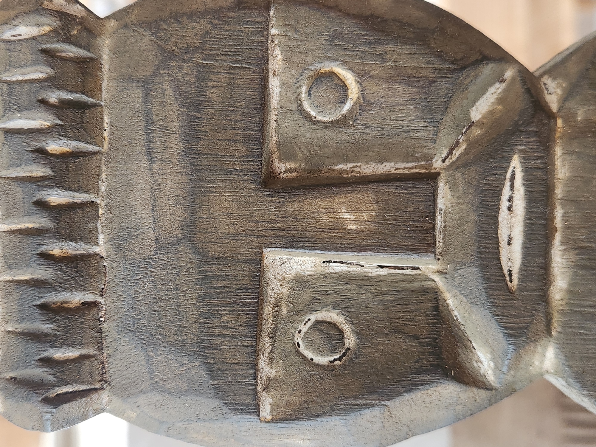 The image shows a close-up, head-on view of a wooden carving of a face, likely an artifact. The carving is dark grey with lighter grey and white accents around the facial features. The top of the carving features a headdress or hair detail with a series of vertical, carved indentations. Below this, the face is broad with a prominent nose. The eyes are depicted as recessed squares, each containing a circular indentation with a smaller, darker circle in the center, resembling pupils. The mouth is a thin, horizontal slit with two small, darker dashes within it. The texture of the wood grain is visible throughout the carving, and there are visible marks and imperfections on its surface. The background is blurred and out of focus, appearing to be a light-colored interior space. There are no people or animals present in the image, and no identifiable landmarks or environmental features that would indicate a specific location within Maitland, United States of America.